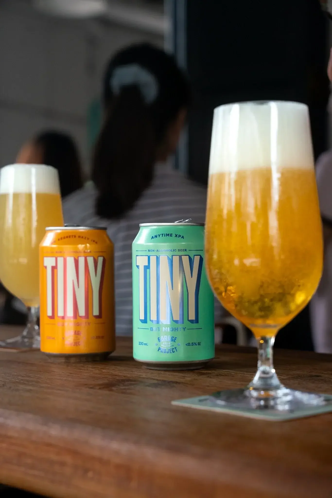 A close-up of cans of non-alcoholic beer and glasses of beer on a wooden table, with a blurred background showing people and a person in striped shirt.