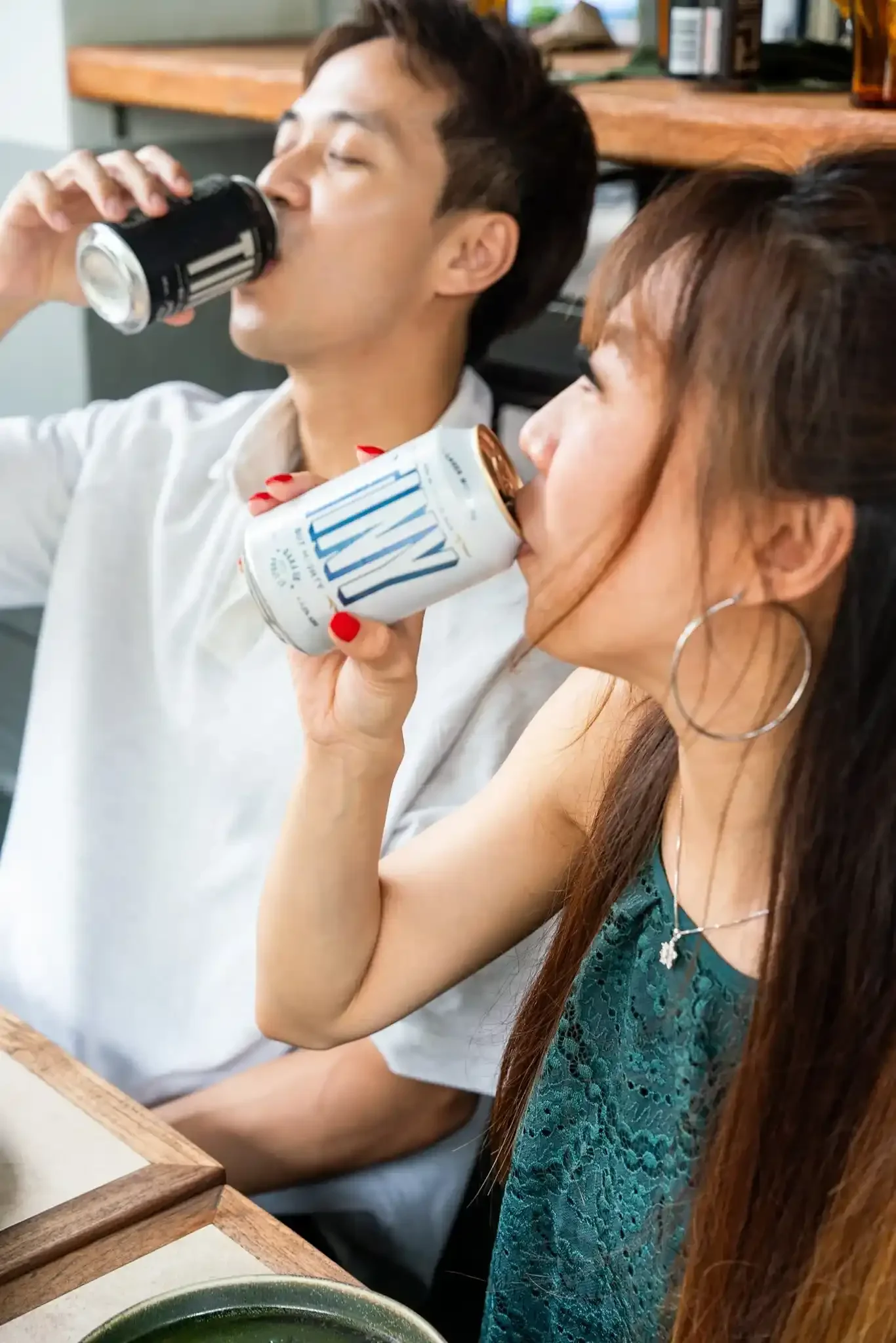 A man drinking TINY Day and Night Stout, and a woman drinking TINY Lager with Lime at a table, sitting close together, with a wooden shelf in the background.