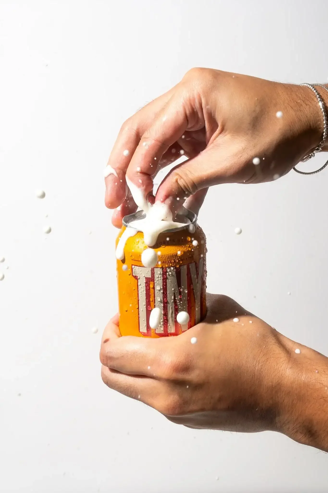 Two hands opening a can of beer, causing foam and liquid to splash out.