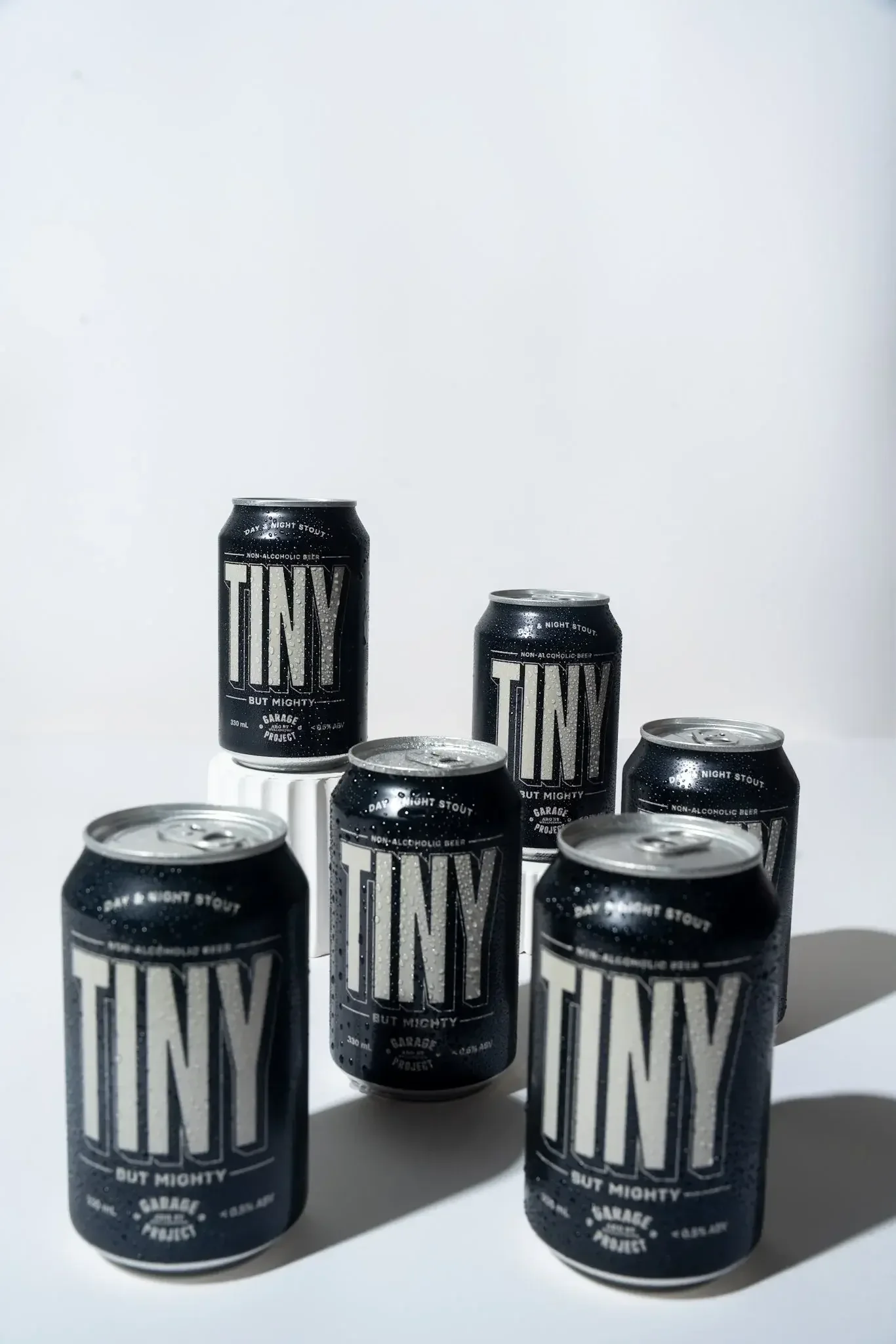 Multiple cans of non-alcoholic stout beer with black and white labels reading "TINY" and "But Mighty" on a white surface against a plain white background.