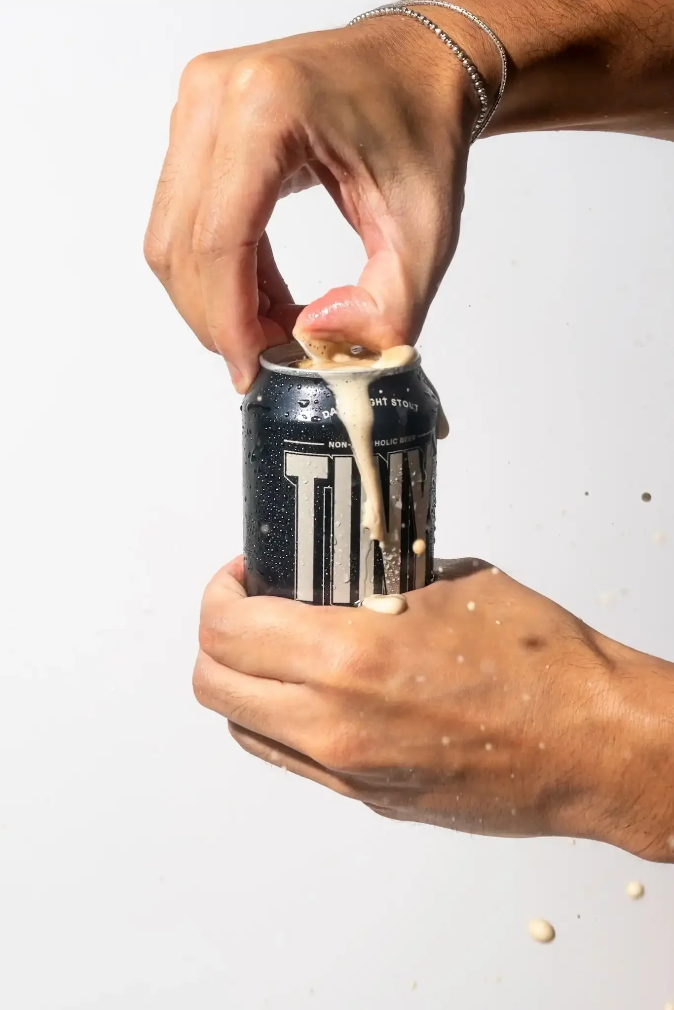 A person's hand is holding a black soda can with white branding, and they are pulling the tab open, causing foam and liquid to spill out.