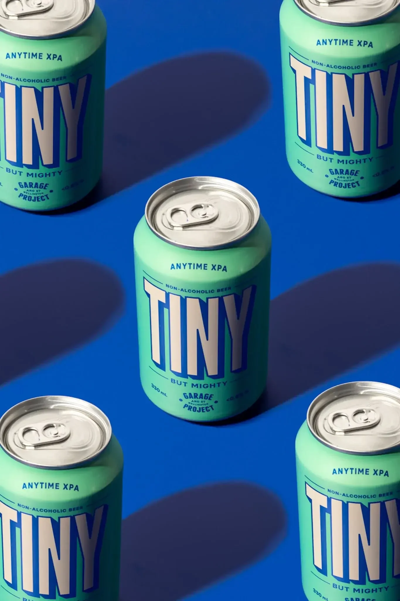 Several cans of non-alcoholic beer called TINY, with a distinct light green color, are arranged on a blue background. The cans have blue and white text and a pull-tab on top.