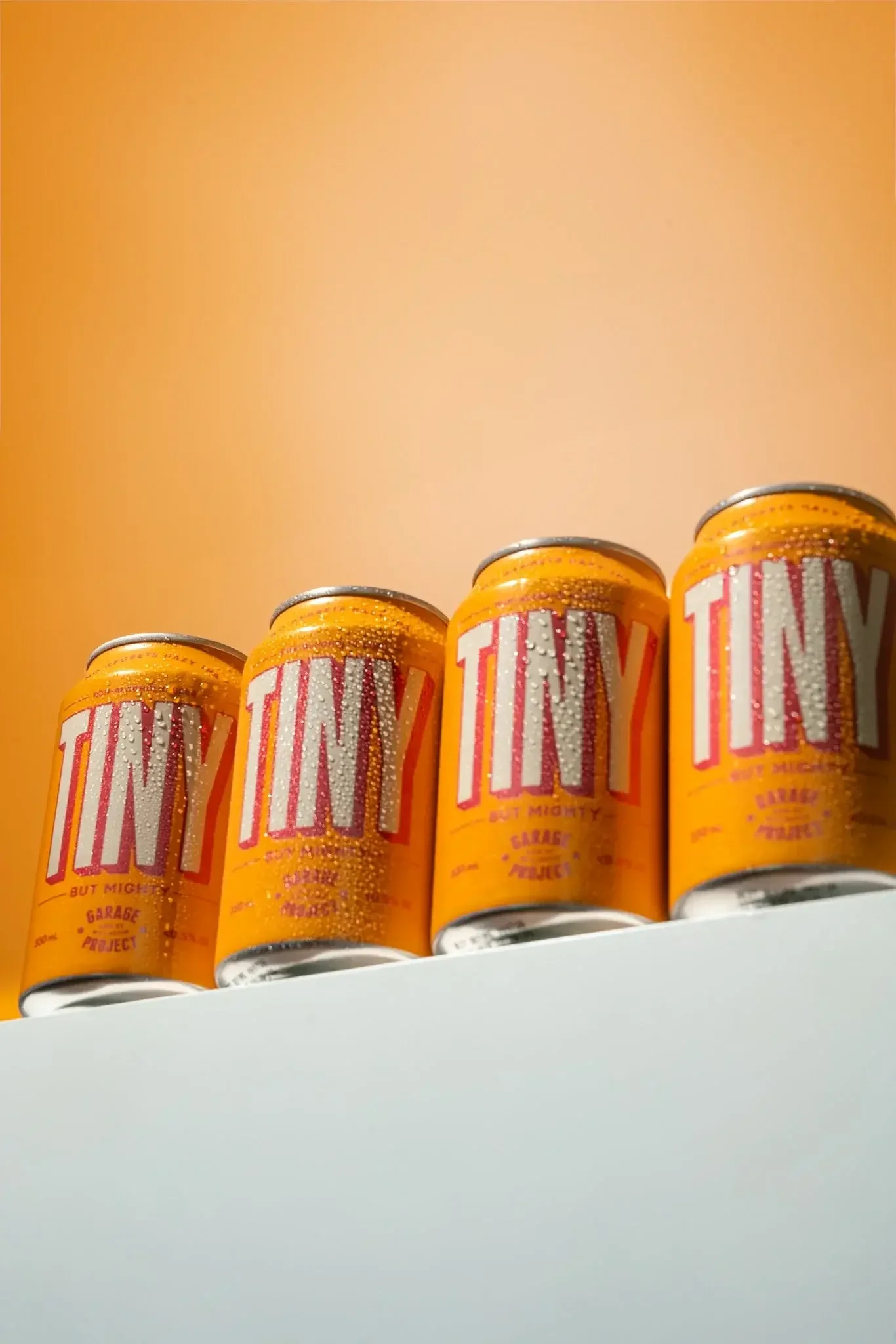 Four cans of orange-colored beverage, labeled 'TINY' with additional text, placed on a white surface against a peach-colored background.