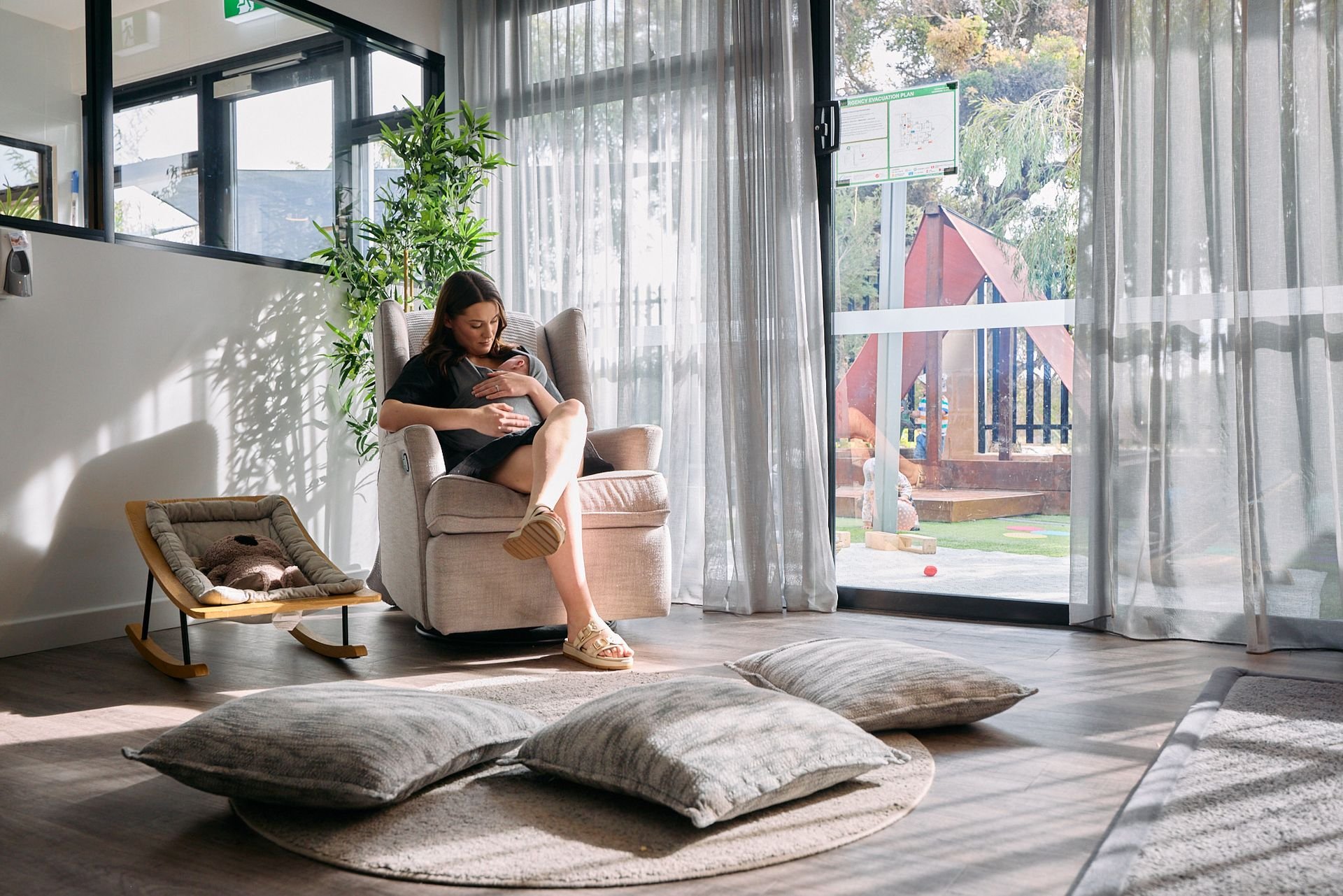 A woman sitting in a beige armchair with her legs crossed, holding her pregnant belly, in a bright living room with sheer curtains and large glass doors leading to an outdoor play area. There is a small pet bed with a sleeping cat on the side, large pillows on the floor, and a tall green plant.