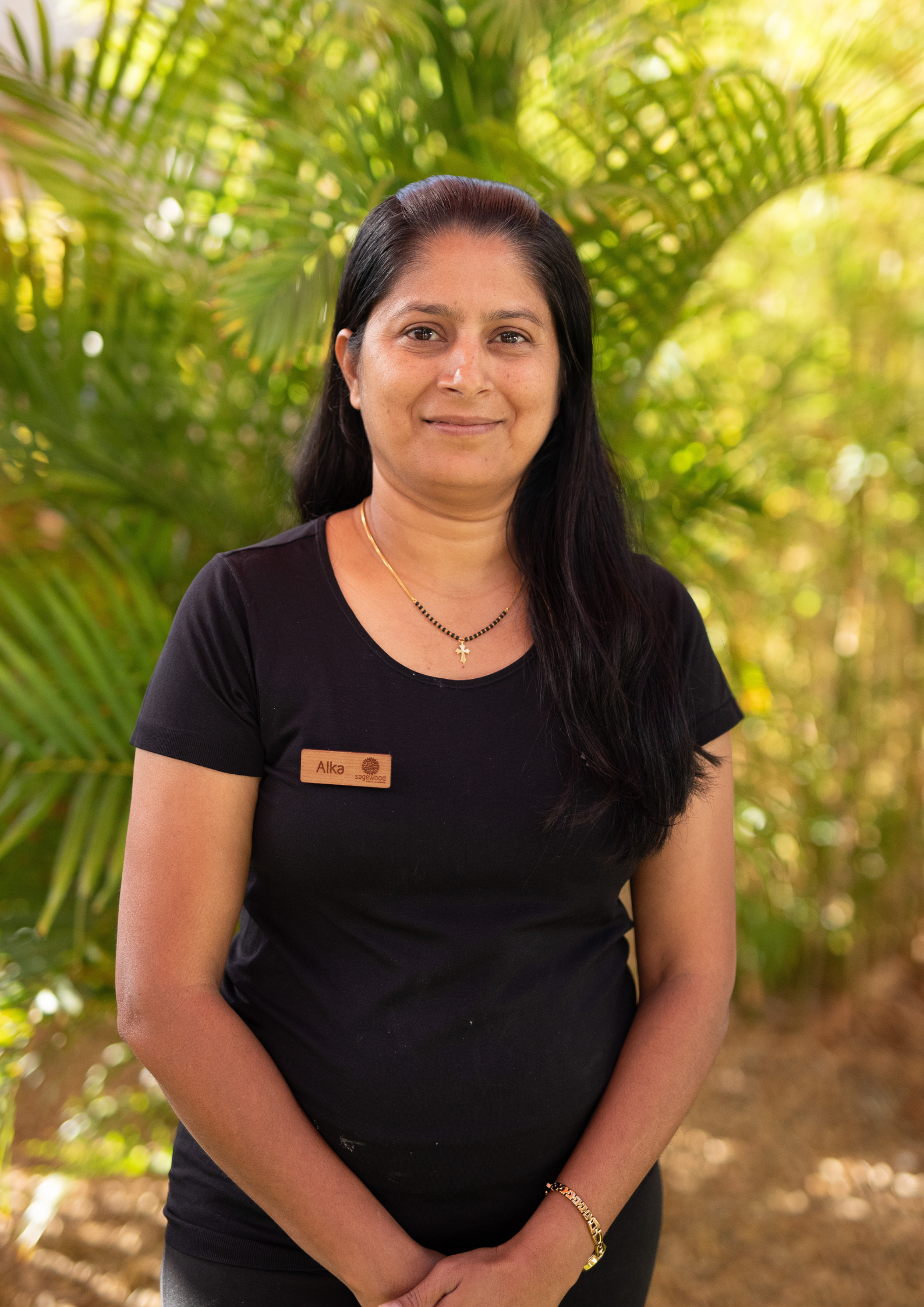 A woman with long black hair wearing a black shirt with a name tag that reads 'Alka' standing outdoors with green foliage in the background.
