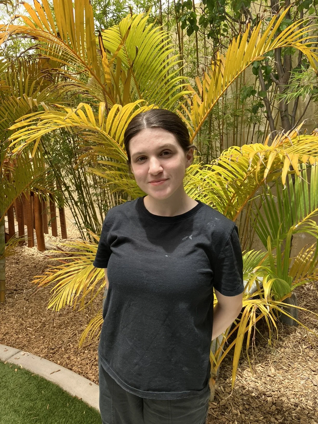 A young woman standing outdoors in front of yellow-green tropical plants, wearing a black T-shirt and dark pants.