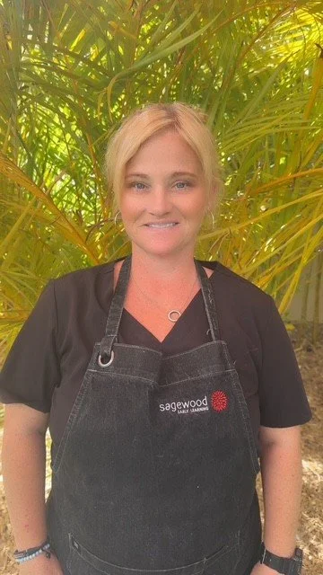 A woman with blonde hair smiling, wearing a black shirt and a black apron with the Sagewood logo, standing outdoors in front of large green palm leaves.