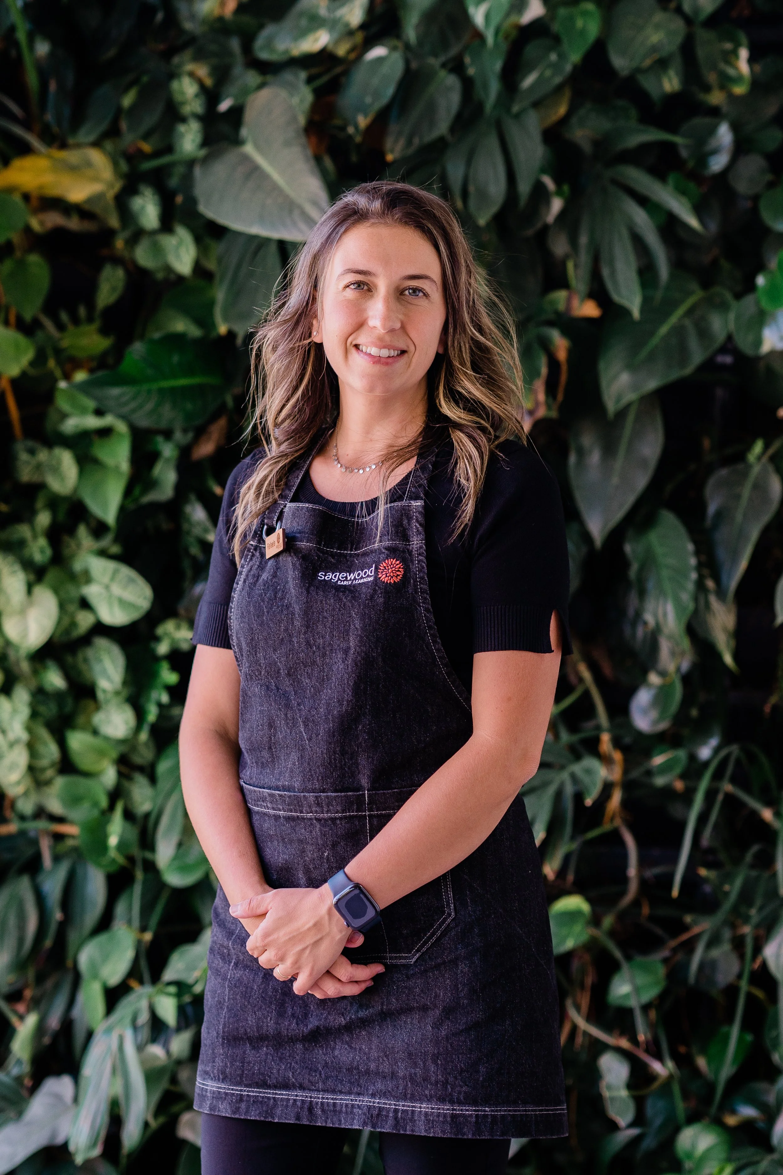 A woman with wavy brown hair wears a black t-shirt, a black apron with 'Sagewood' embroidered on it, and a smartwatch, standing in front of a lush green leafy background.