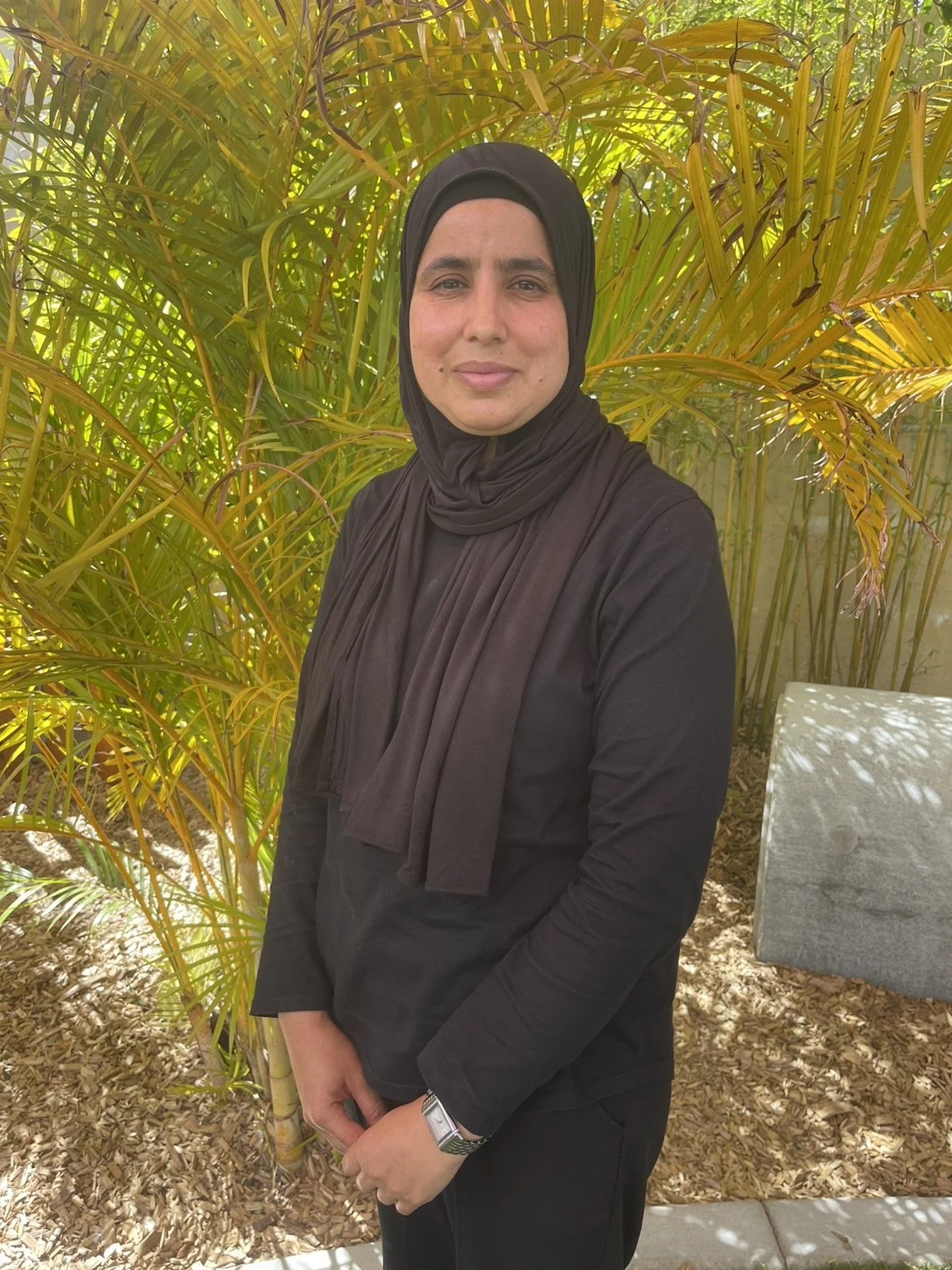 A woman wearing a black hijab and black clothing stands outdoors in front of green and yellow plants, smiling softly at the camera.