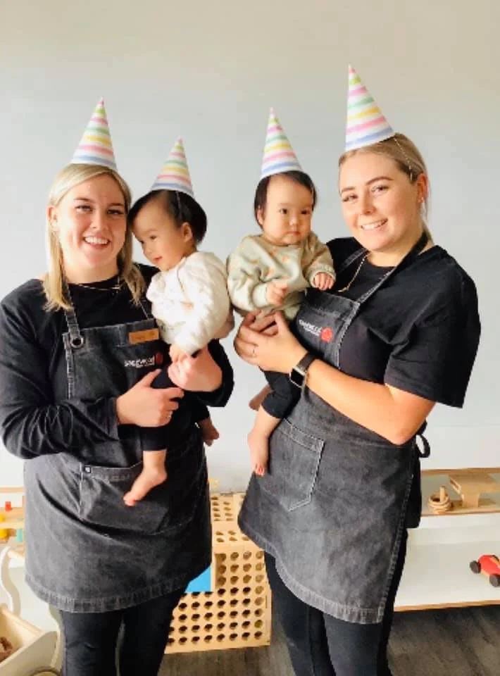Two women and two young children wearing birthday party hats, smiling and posing for a photo indoors.