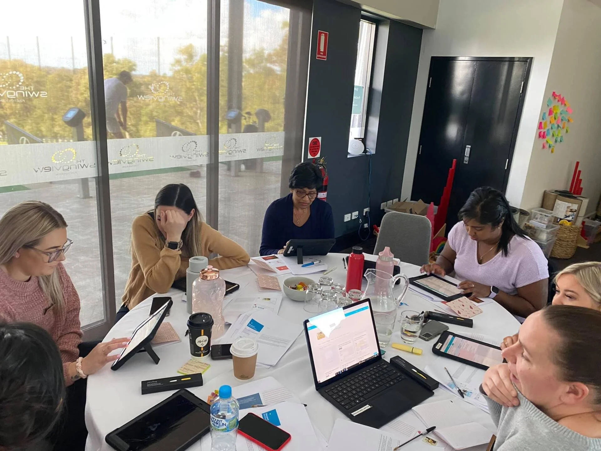 A group of women sitting around a white conference table, using laptops and tablets in a meeting room with large windows showing an outdoor exercise area.