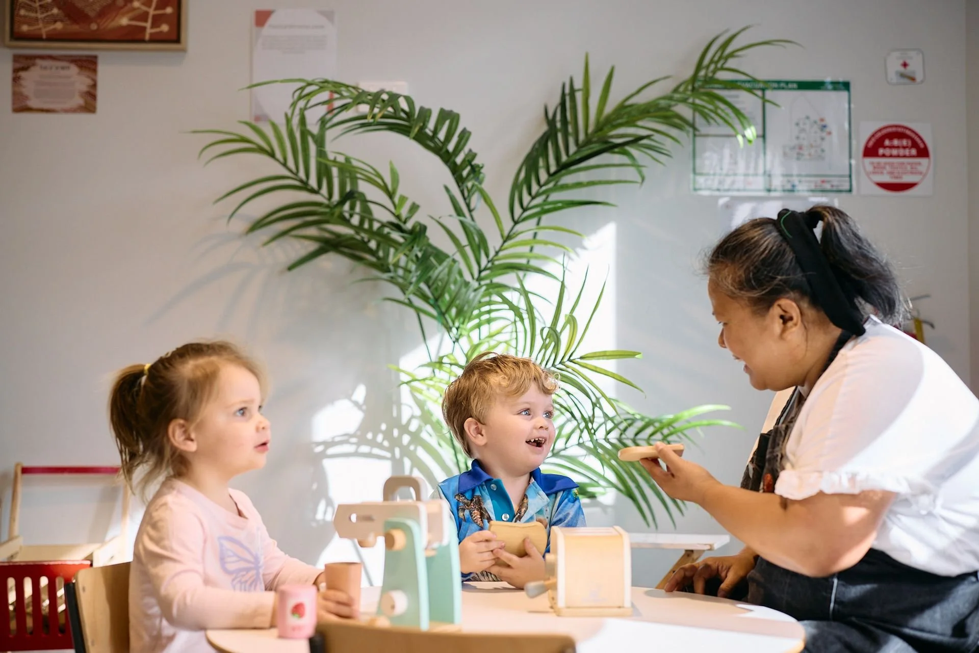 A caregiver in a white shirt and black apron smiles at two young children sitting at a table in a room with a large green plant in the background. The children are playing with wooden toy cash register and other toys, and the room has bright natural light.