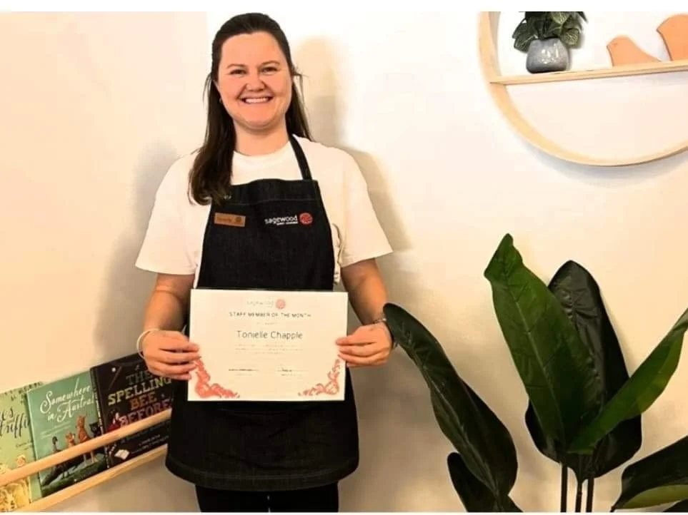 A woman smiling and holding a certificate, standing indoors next to a potted plant and books on a shelf.