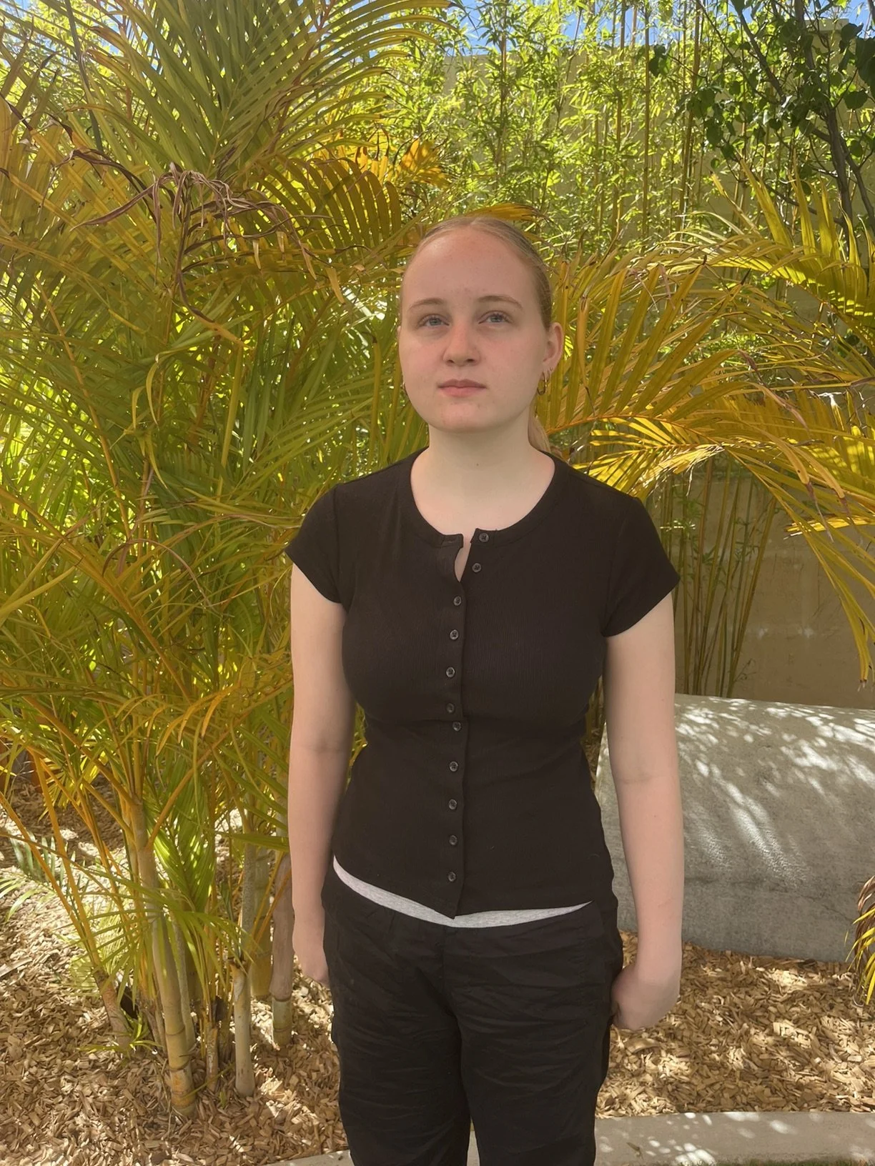 A young woman in a black shirt and black pants standing outdoors in front of lush green plants.