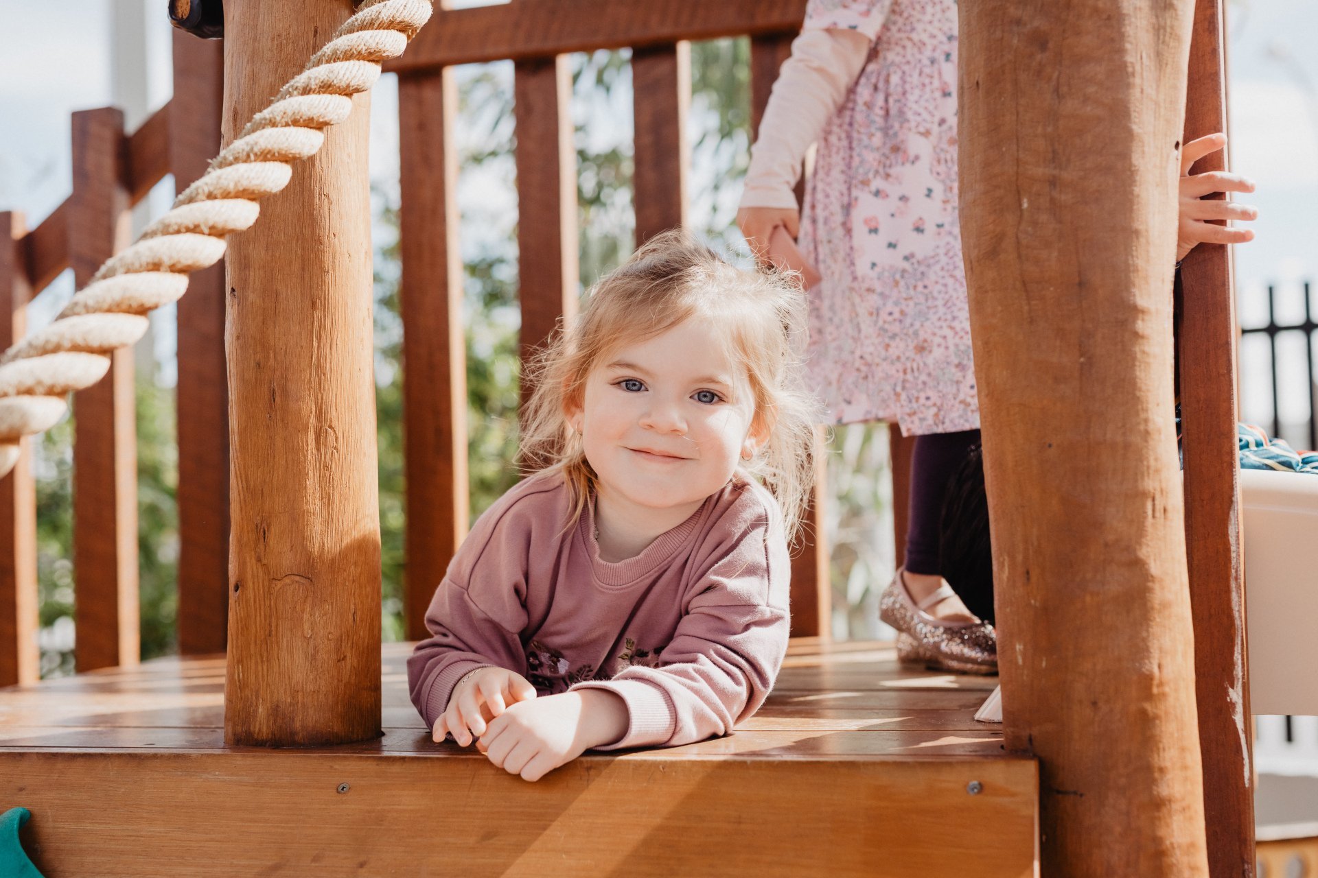 A young girl with blonde hair and blue eyes, lying on her stomach on a wooden deck, smiling. Another child, partially visible, is standing behind her on the deck.