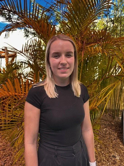 A young woman standing outdoors in front of tropical plants with green and orange fronds, wearing a black t-shirt.