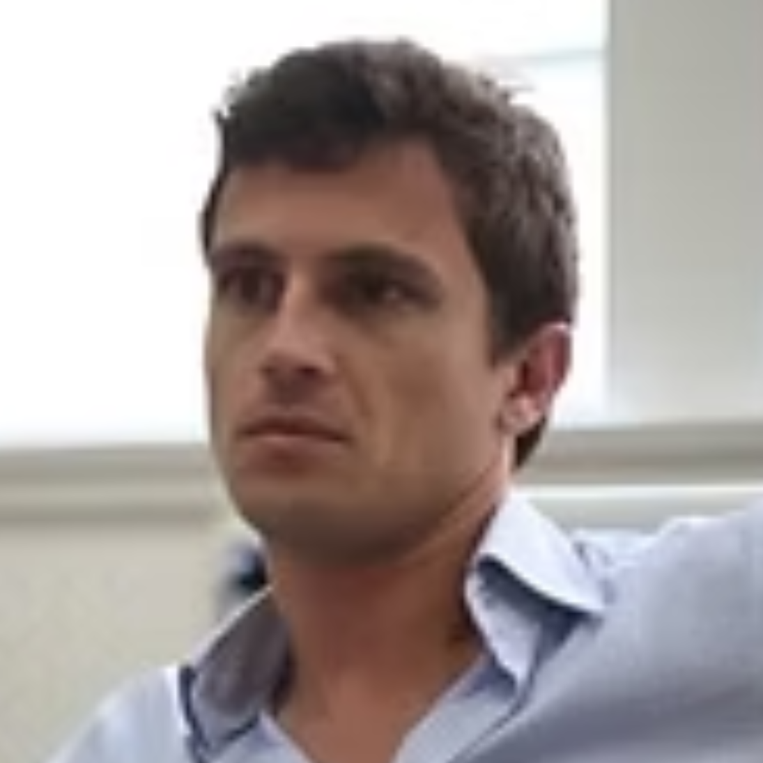 A young man with dark hair wearing a light blue collared shirt, looking to the side, in an indoor setting with windows in the background.