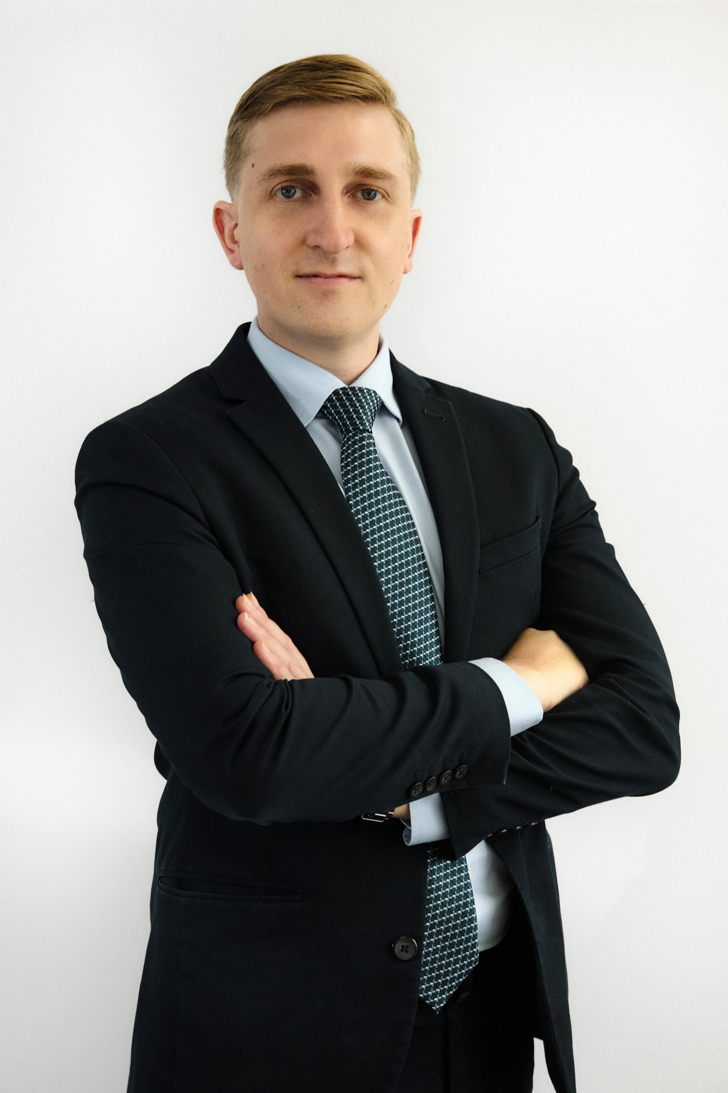 A young man with blonde hair and blue eyes in a suit, standing with arms crossed against a plain white background.
