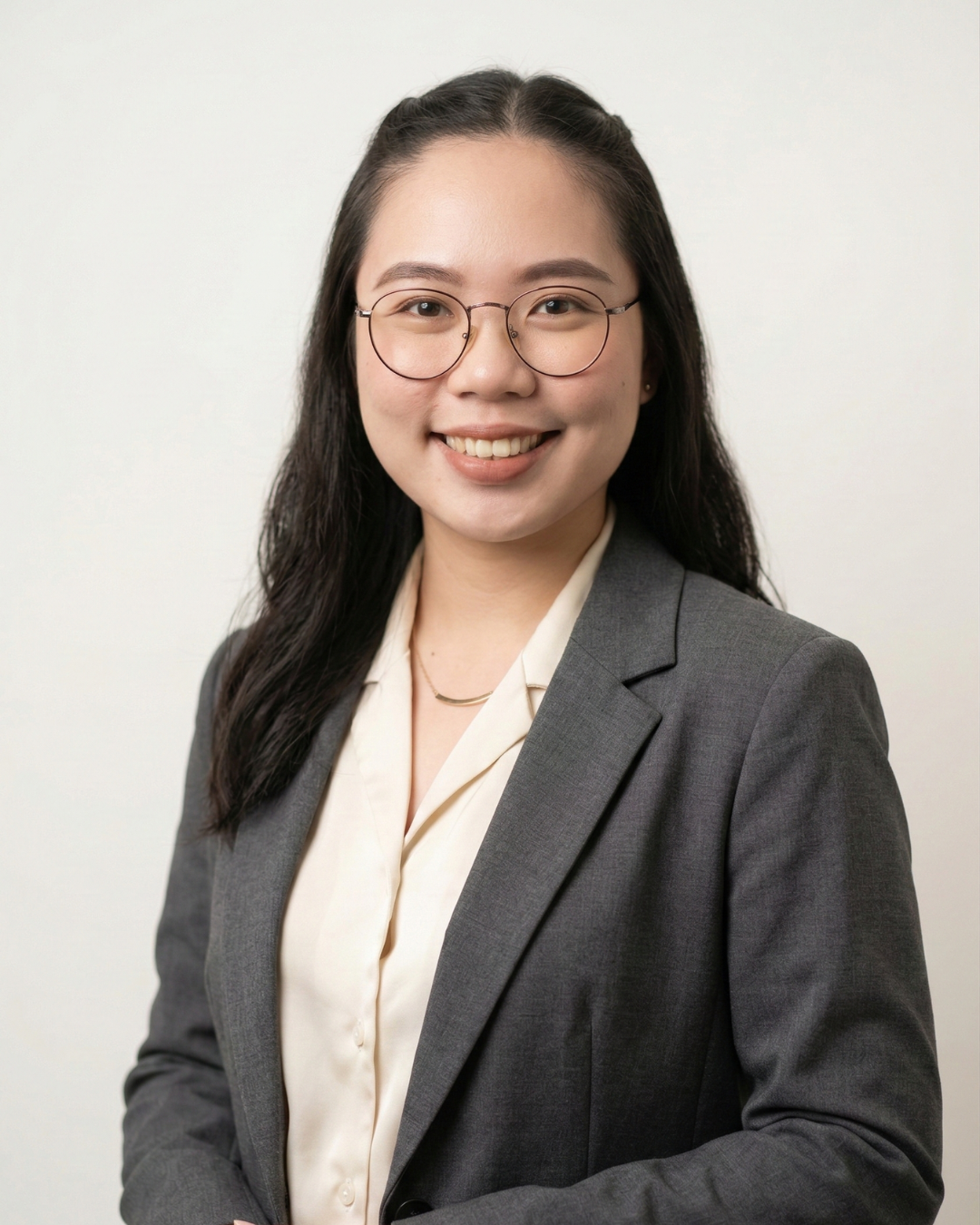 A young woman with long black hair and glasses, wearing a gray blazer and cream blouse, smiling confidently against a plain off-white background.