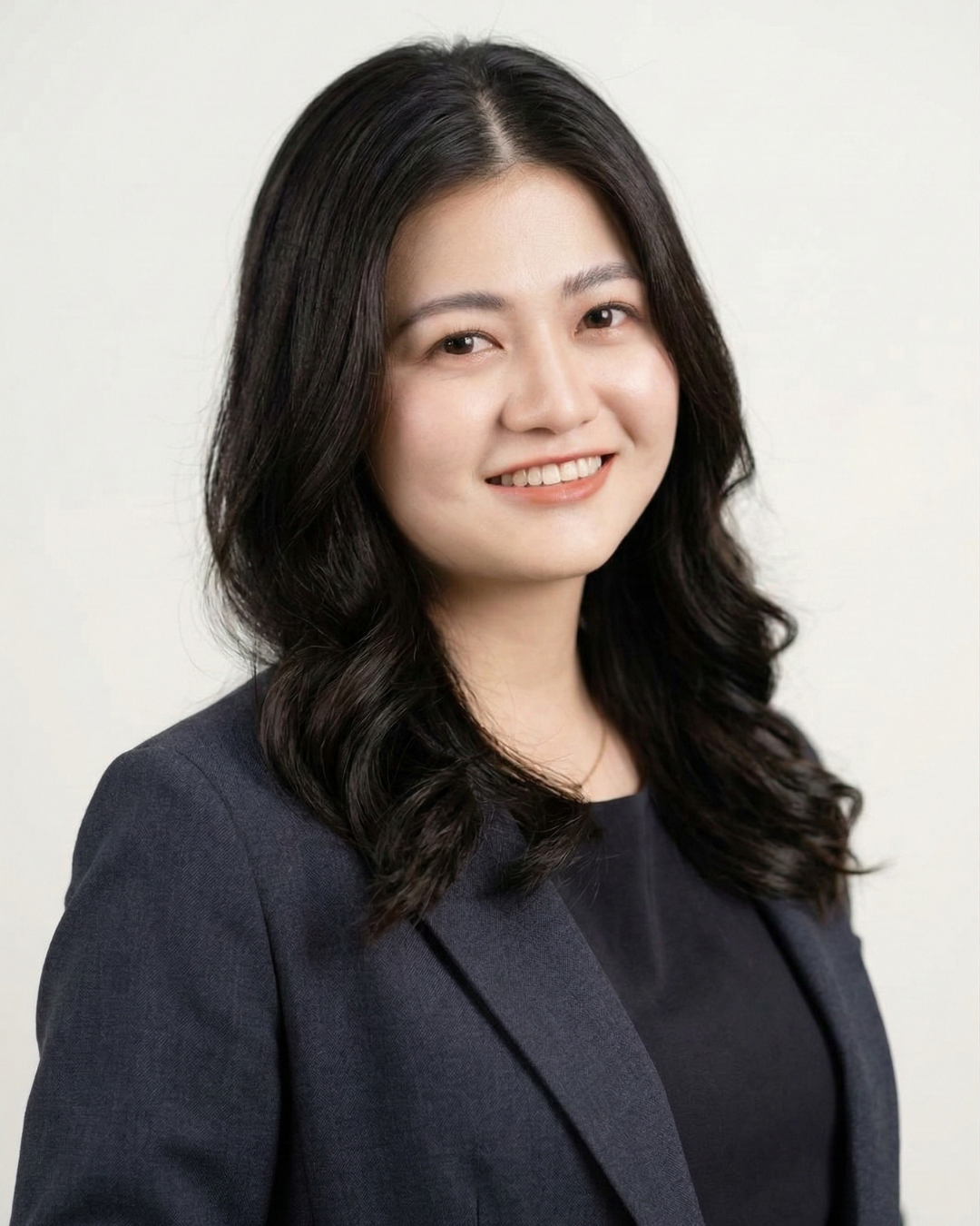 Portrait of a young woman with long black hair, smiling, wearing a dark blazer and black top, against a plain white background.