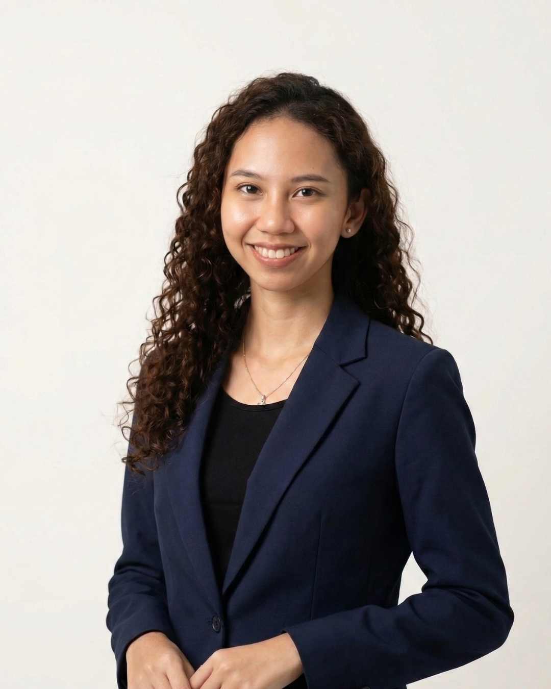 A young woman with curly hair, wearing a navy blazer over a black top, smiling, standing against a plain white background.