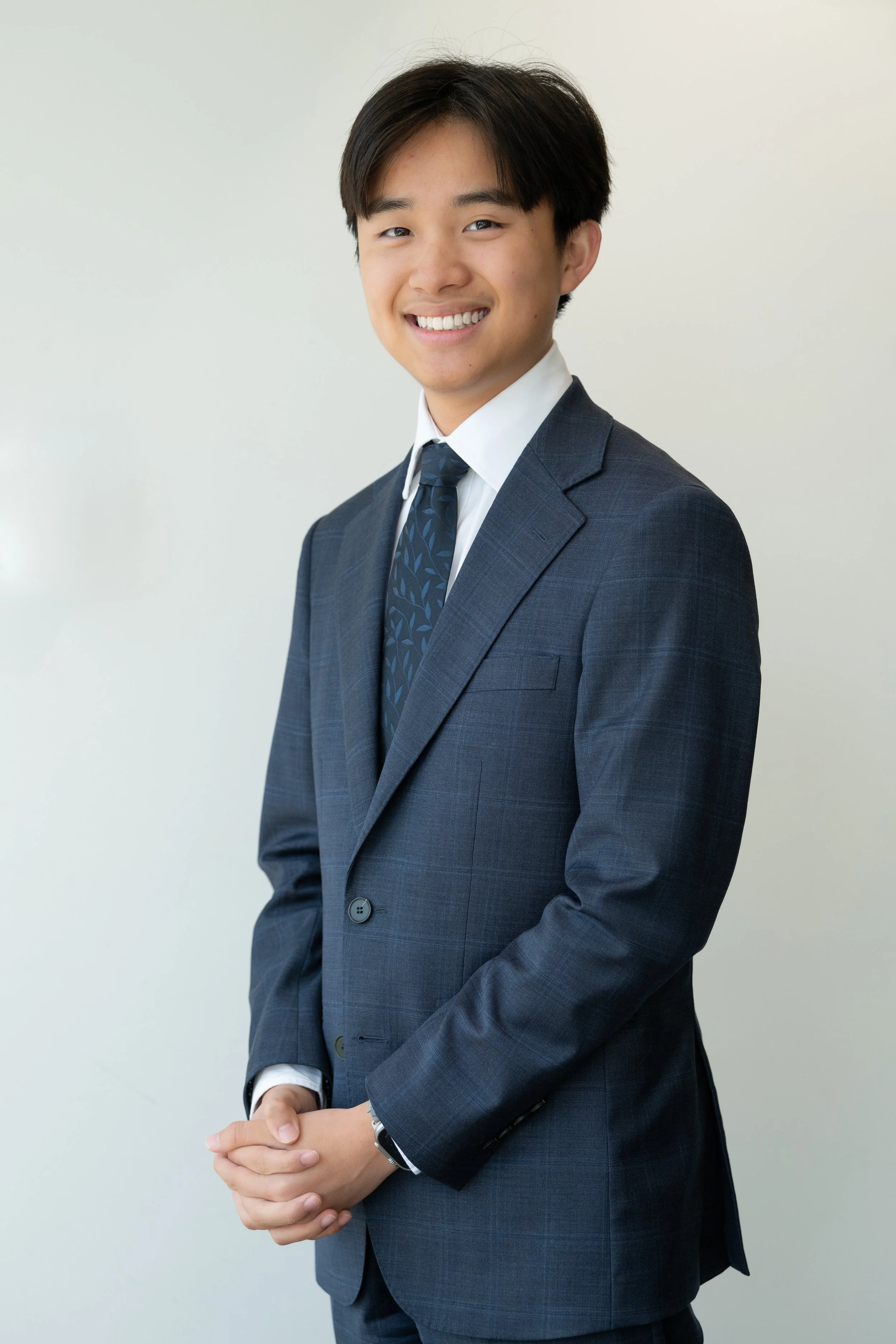 A young man in a dark blue suit, white shirt, and blue patterned tie standing against a plain light-colored background, smiling with hands clasped in front.