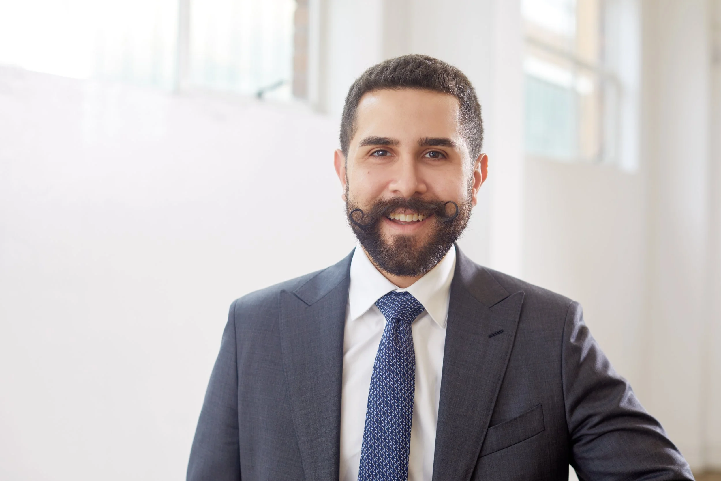 A man with a neatly styled hairstyle and a trimmed beard, wearing a suit, white shirt, and patterned tie, smiling in front of a bright window.