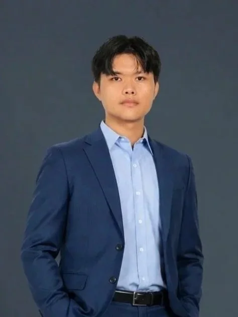 A young man wearing a dark blue suit and light blue dress shirt standing against a dark gray background.