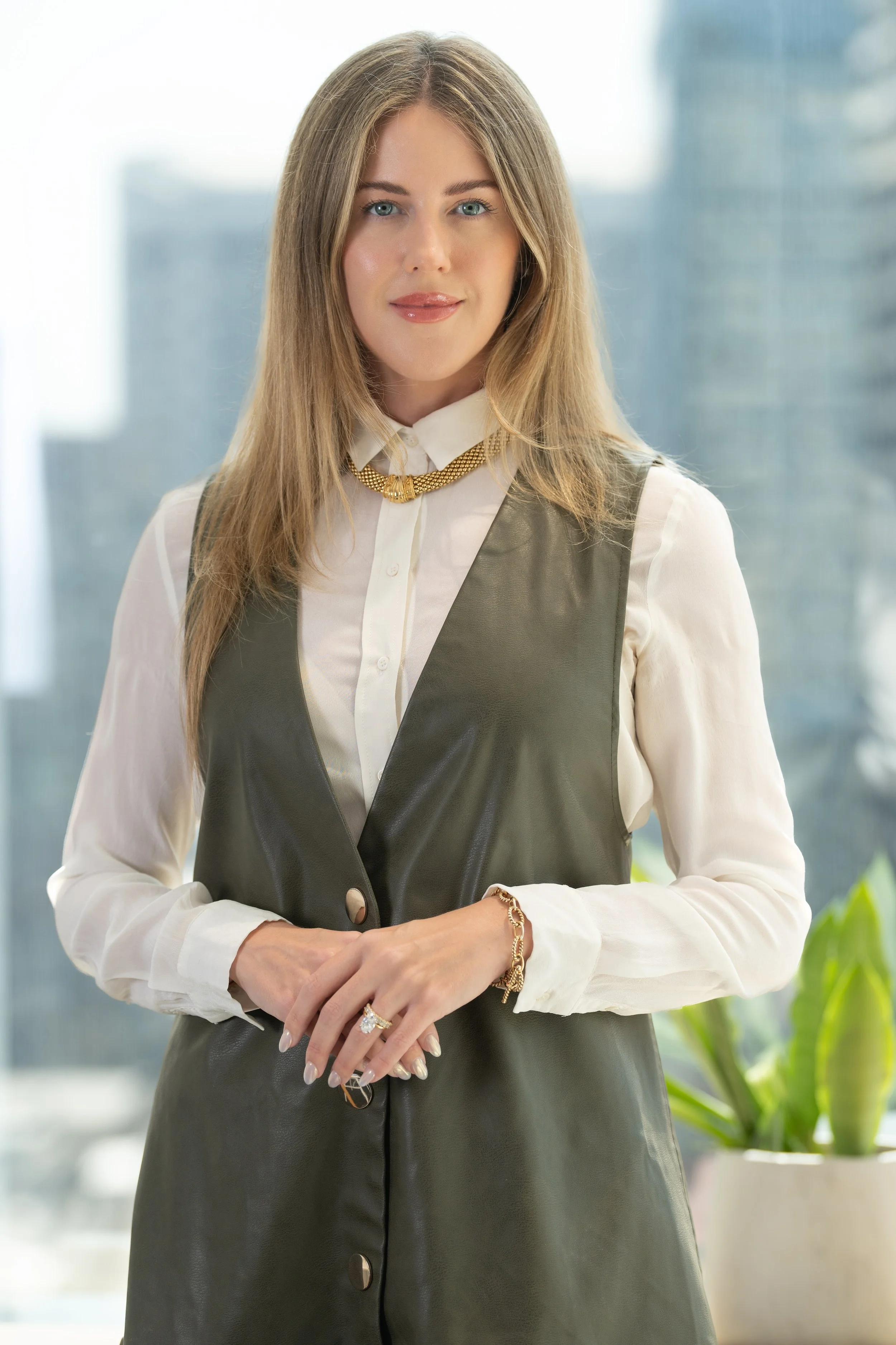 A woman with long blonde hair wearing a white blouse, gray vest, and gold jewelry standing in a modern office setting.