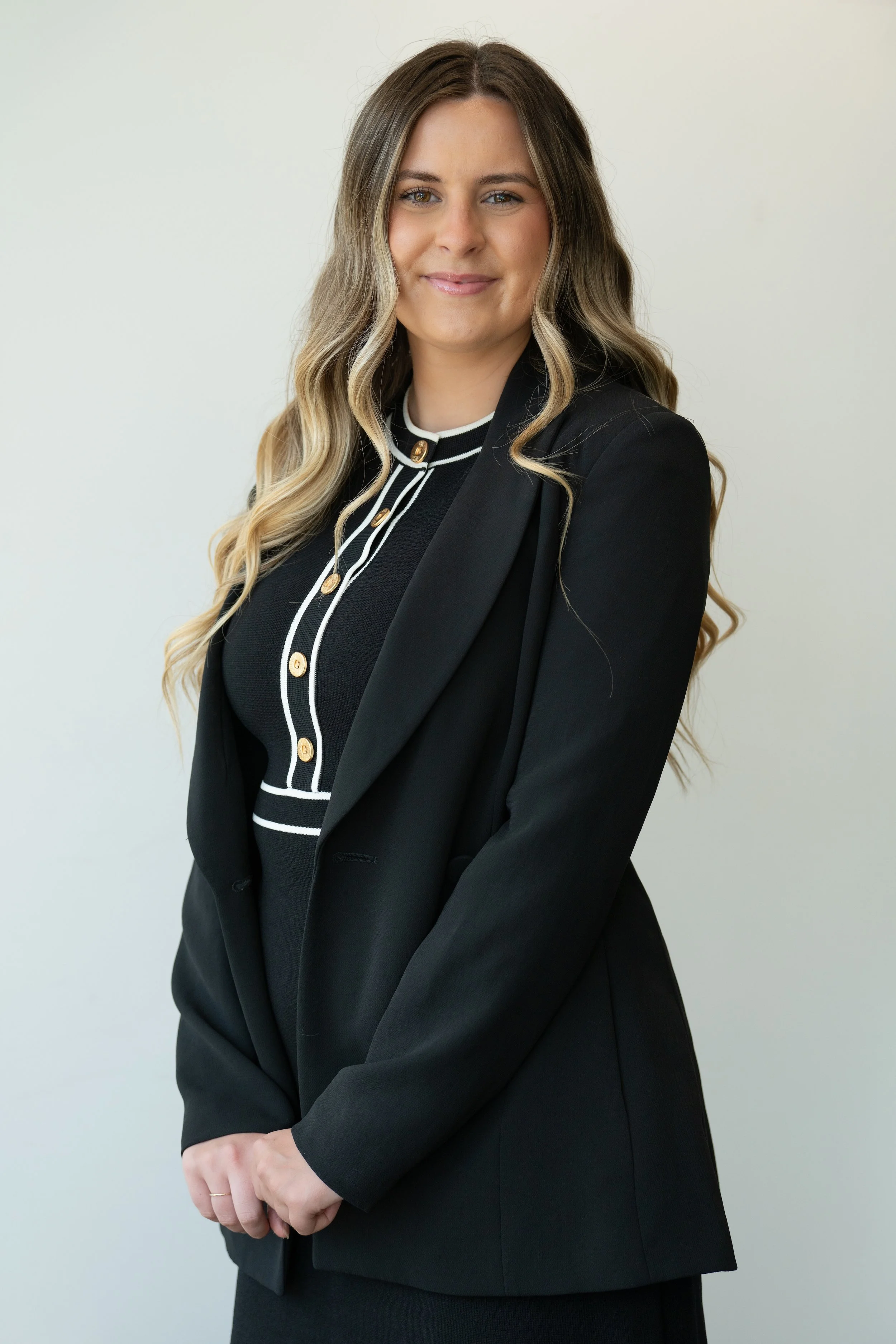 A professional woman with long, wavy blonde hair and light skin, wearing a black blazer and a black top with white trim and gold buttons, standing against a plain light-colored background, smiling softly.