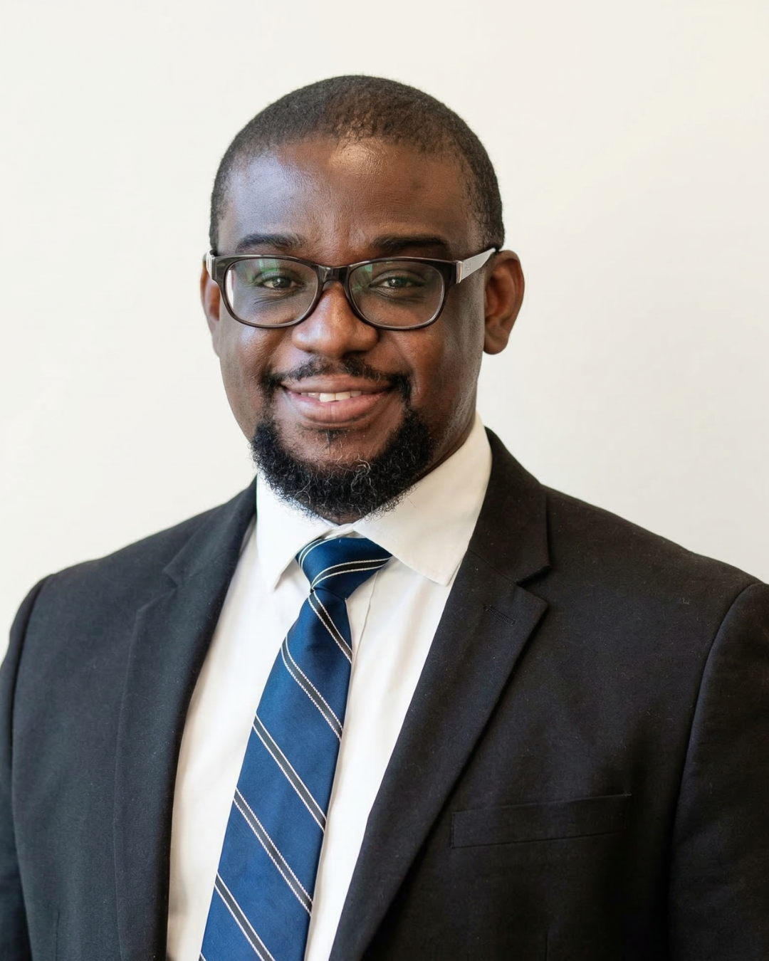 Professional portrait of a smiling African American man wearing glasses, a dark suit, white shirt, and a blue striped tie, against a plain white background.
