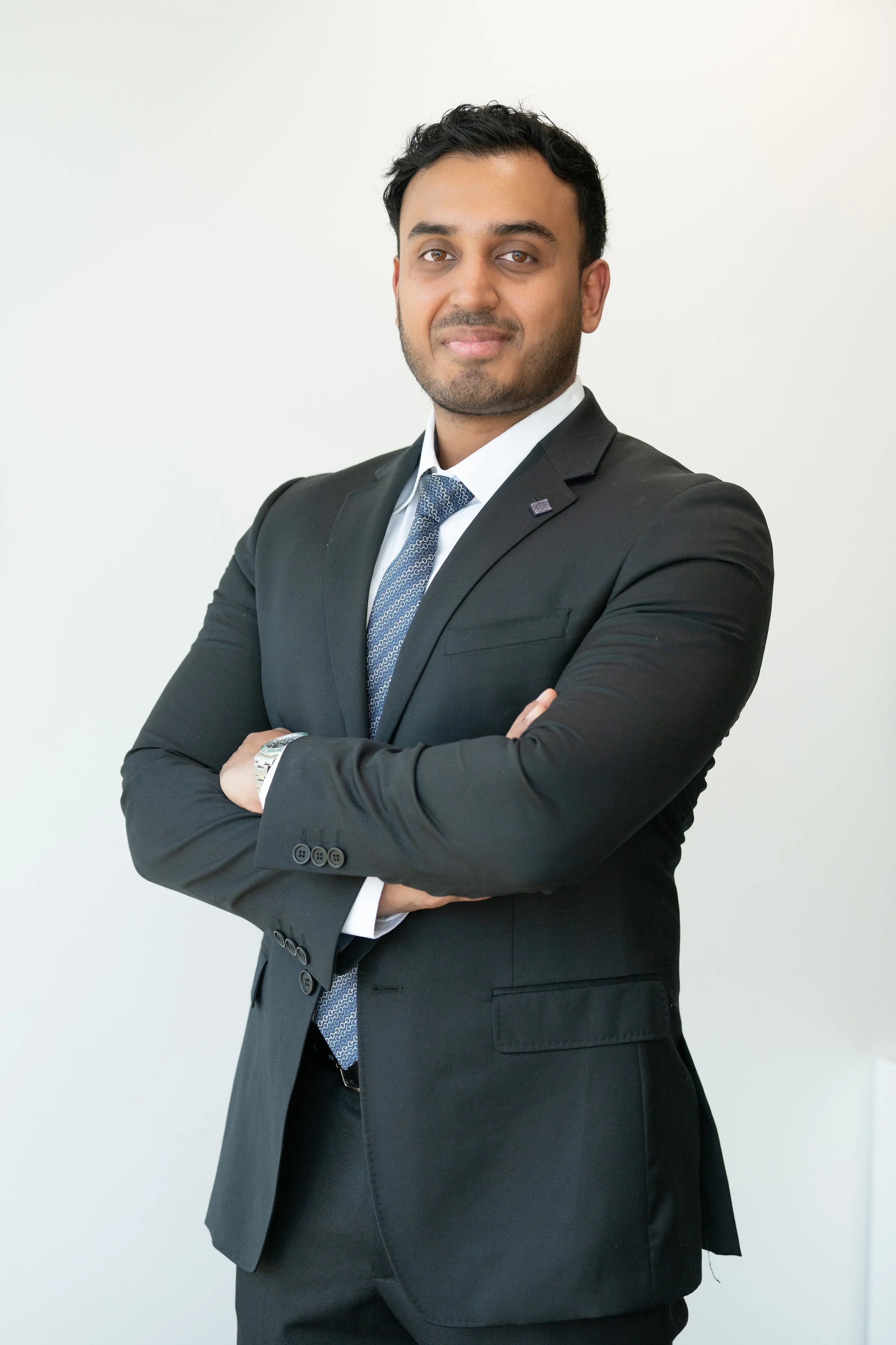 A confident man in a black suit with crossed arms, standing against a plain white background.