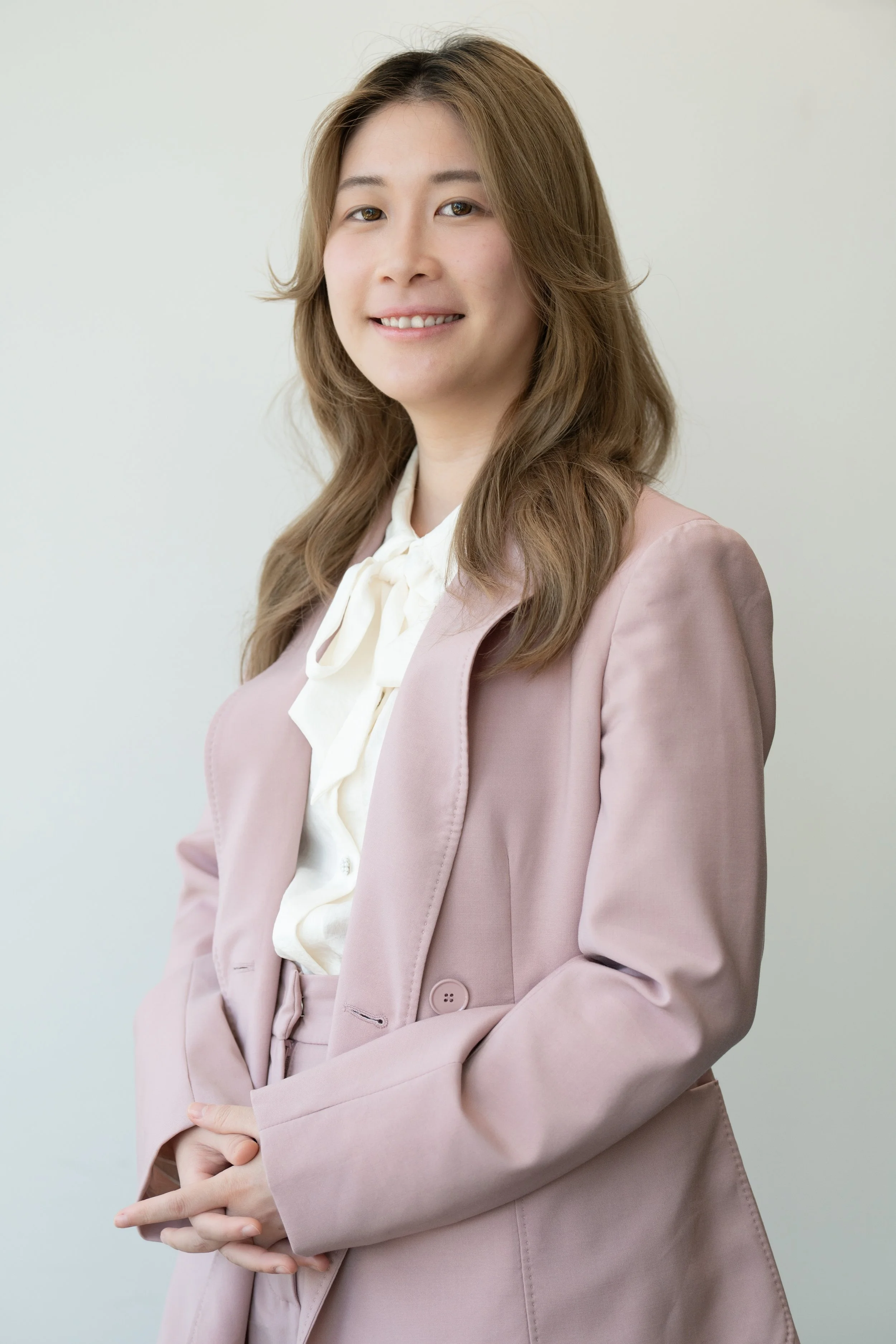 A woman in a light pink suit with a cream blouse, standing with her hands clasped in front of her, smiling gently at the camera, against a plain, off-white background.