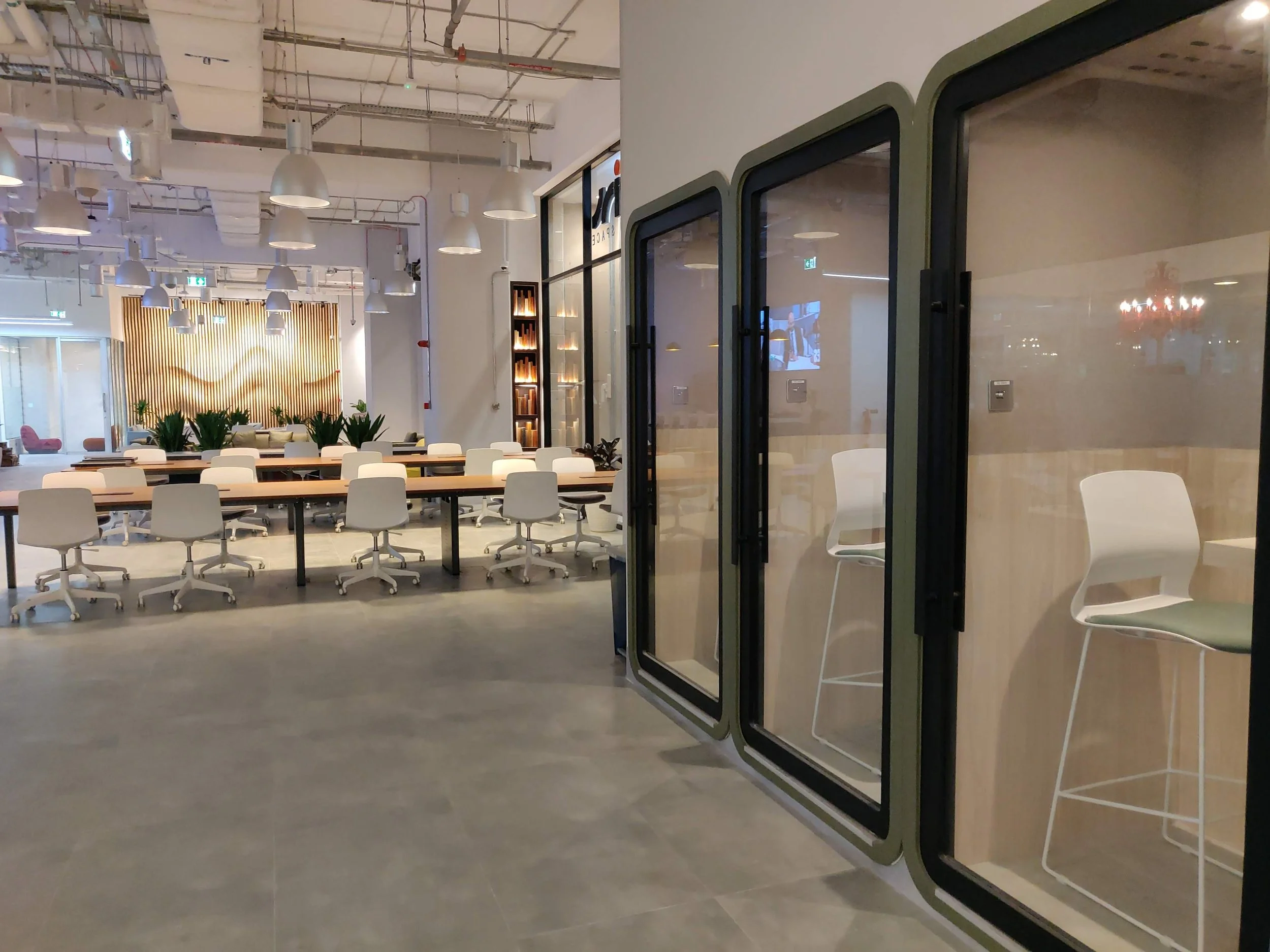 Empty modern office space with glass enclosed work pods on the right, a long table with white chairs at the center, and a lounge area with pink chairs and a wooden wall with plants in the background.