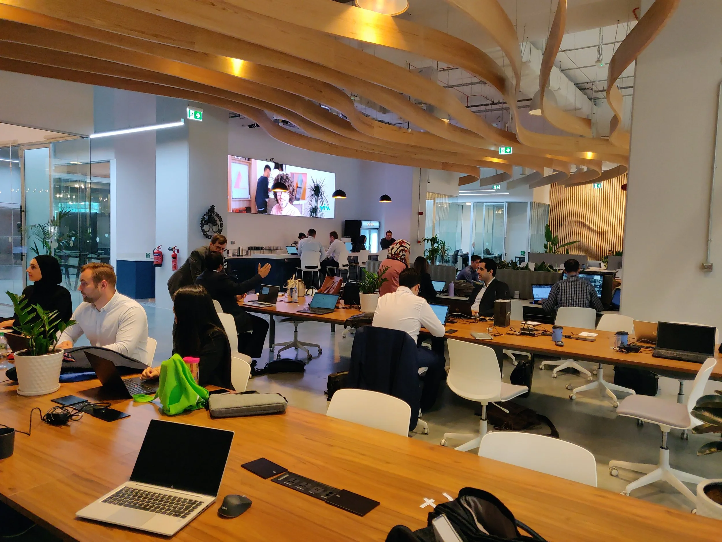 An open-concept coworking space with people working at tables, laptops, and office supplies. There are wooden ceiling accents, indoor plants, and a large digital screen on the wall.