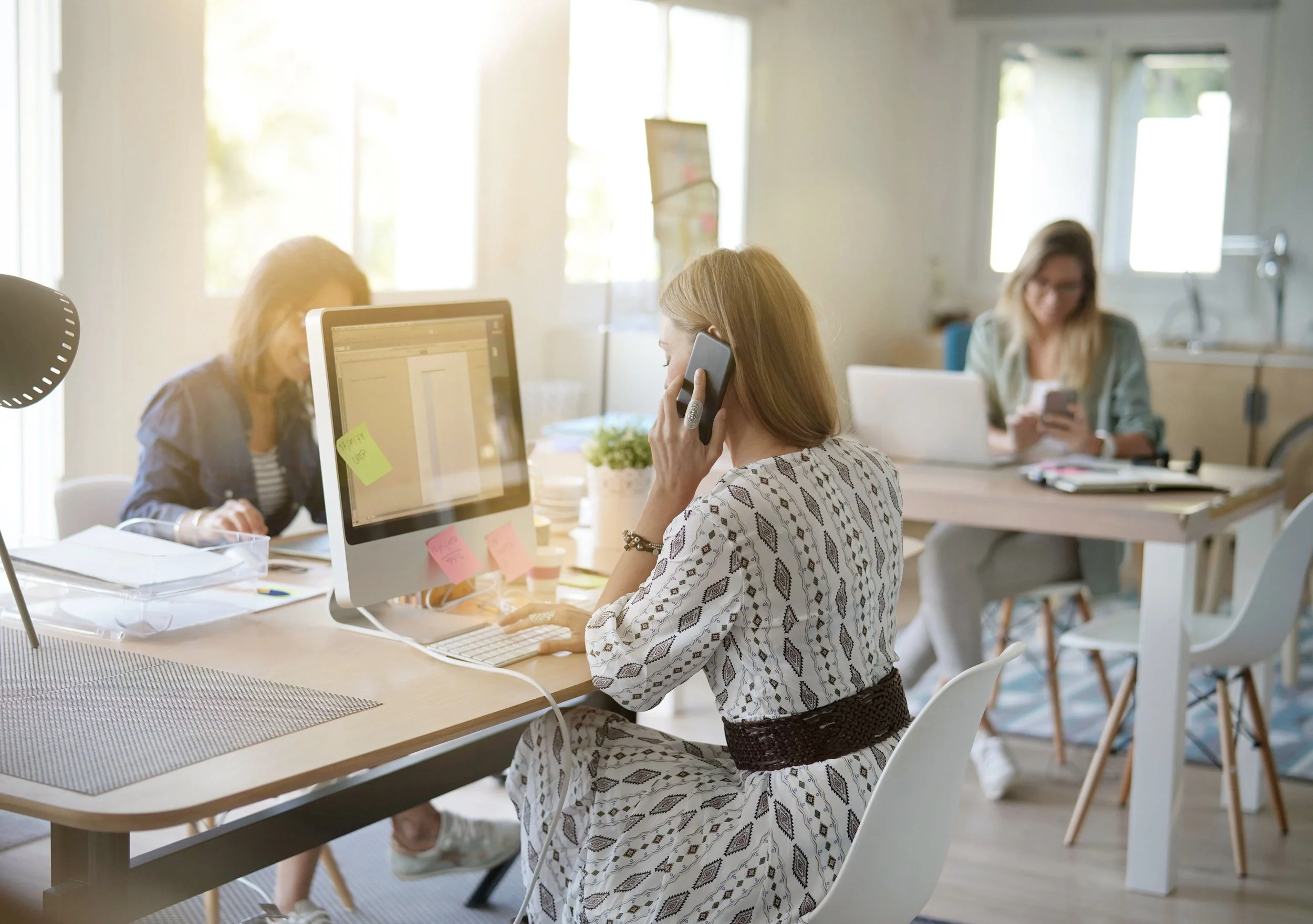 Women working in a modern office with computers, notebooks, and phones, with sunlight coming through large windows.