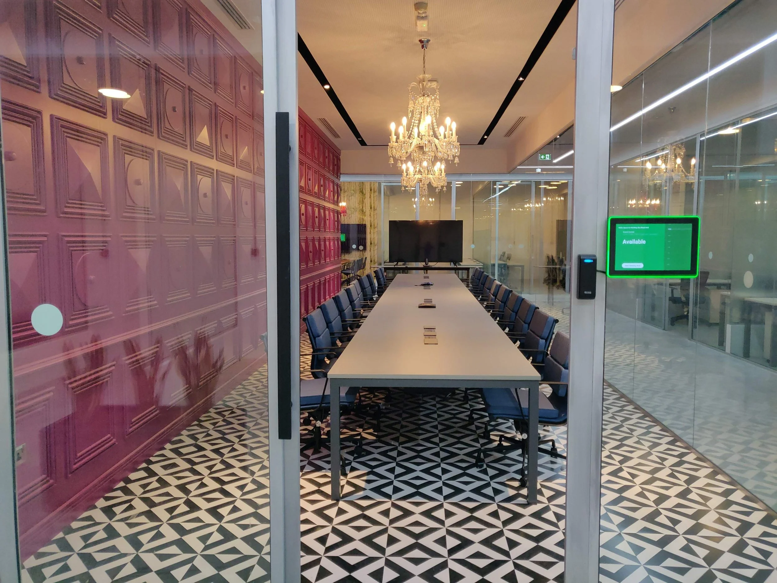 A modern conference room viewed through glass walls, featuring a long table, blue chairs, a chandelier hanging from the ceiling, a large TV screen at the far end, and a patterned black-and-white floor.