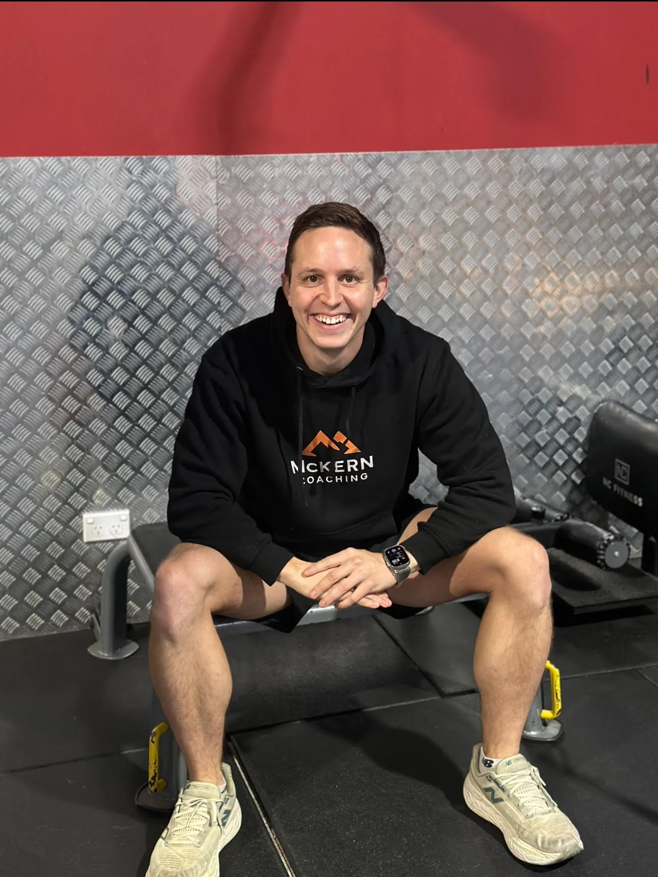 A smiling man in a black hoodie with 'McKern Coaching' logo, sitting on a workout bench, wearing beige sneakers and a smartwatch, in a gym with metallic walls.