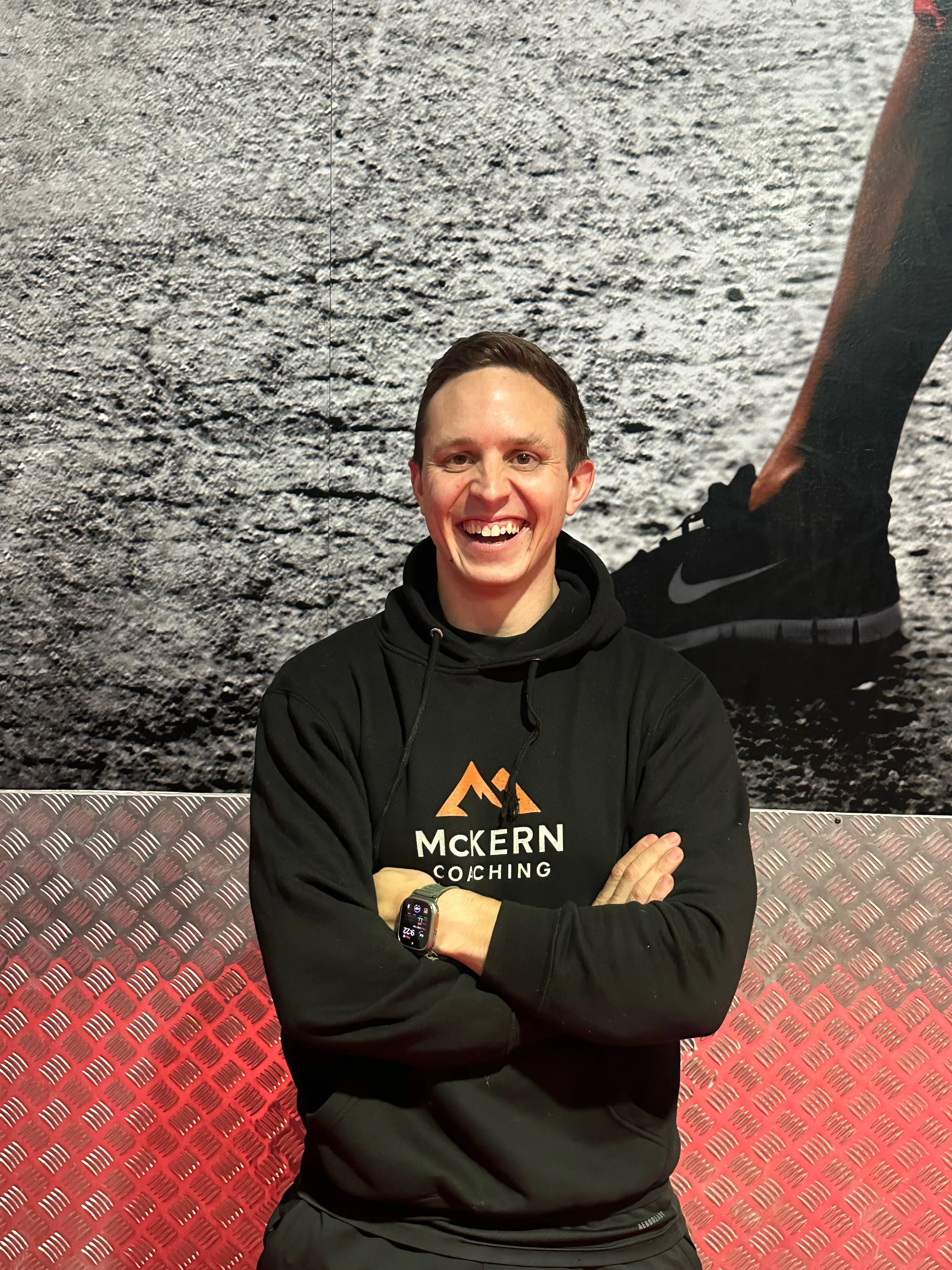 A man in a black hoodie smiling, standing with arms crossed in front of a wall with a textured gray and red surface, and a large poster of a person running, wearing black Nike shoes.