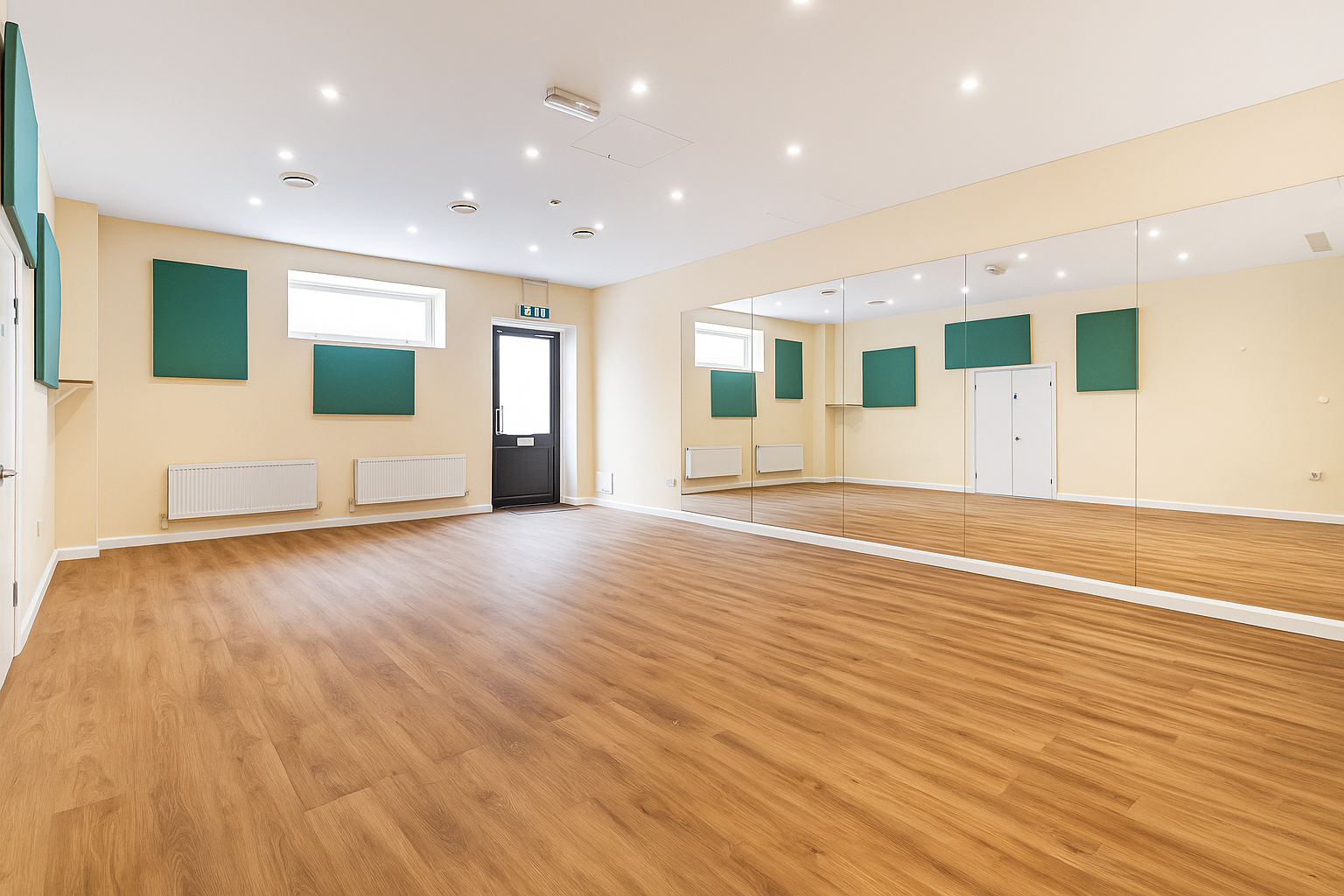 Empty dance studio with wooden floor, cream walls, green acoustic panels, large mirror, black door, and small windows.