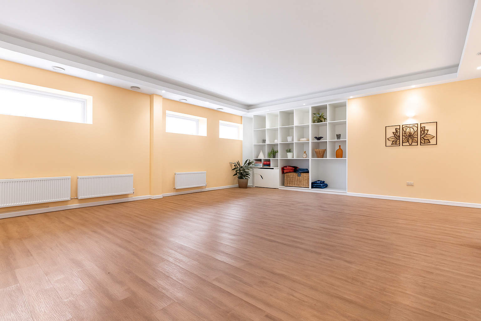 Empty room with wooden floors, yellow walls, white shelving unit with decorative items, potted plant, and three small windows near ceiling.