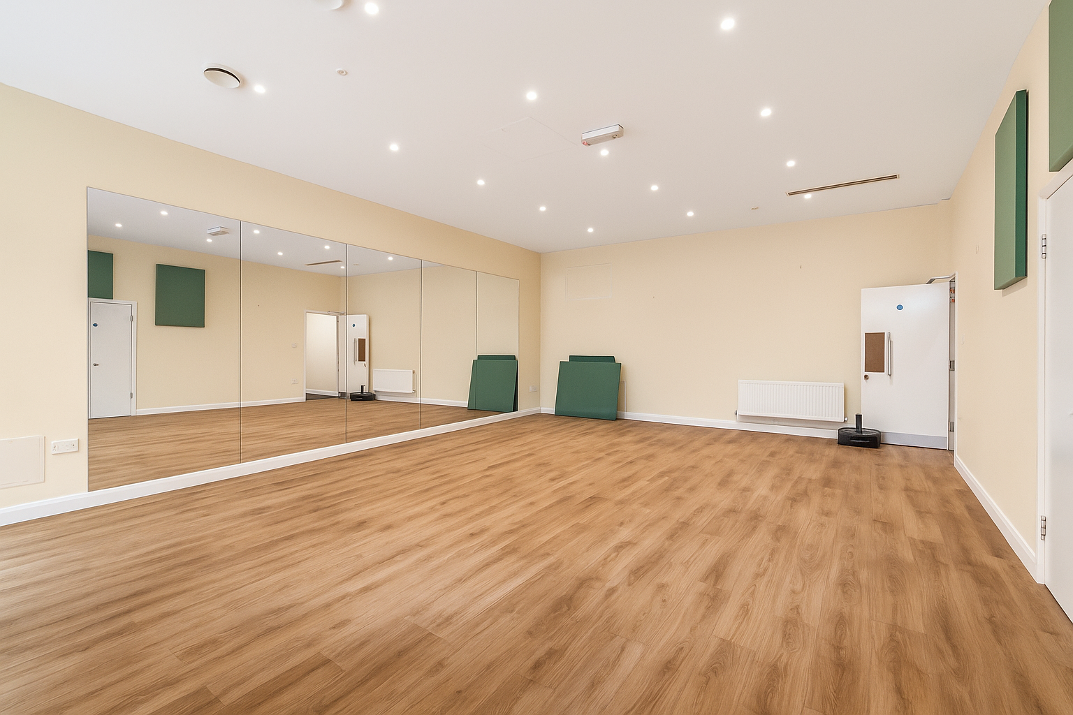 Empty dance or exercise studio with wooden floors, a large mirror wall, white walls, green mats stacked in corners, and ceiling lights.