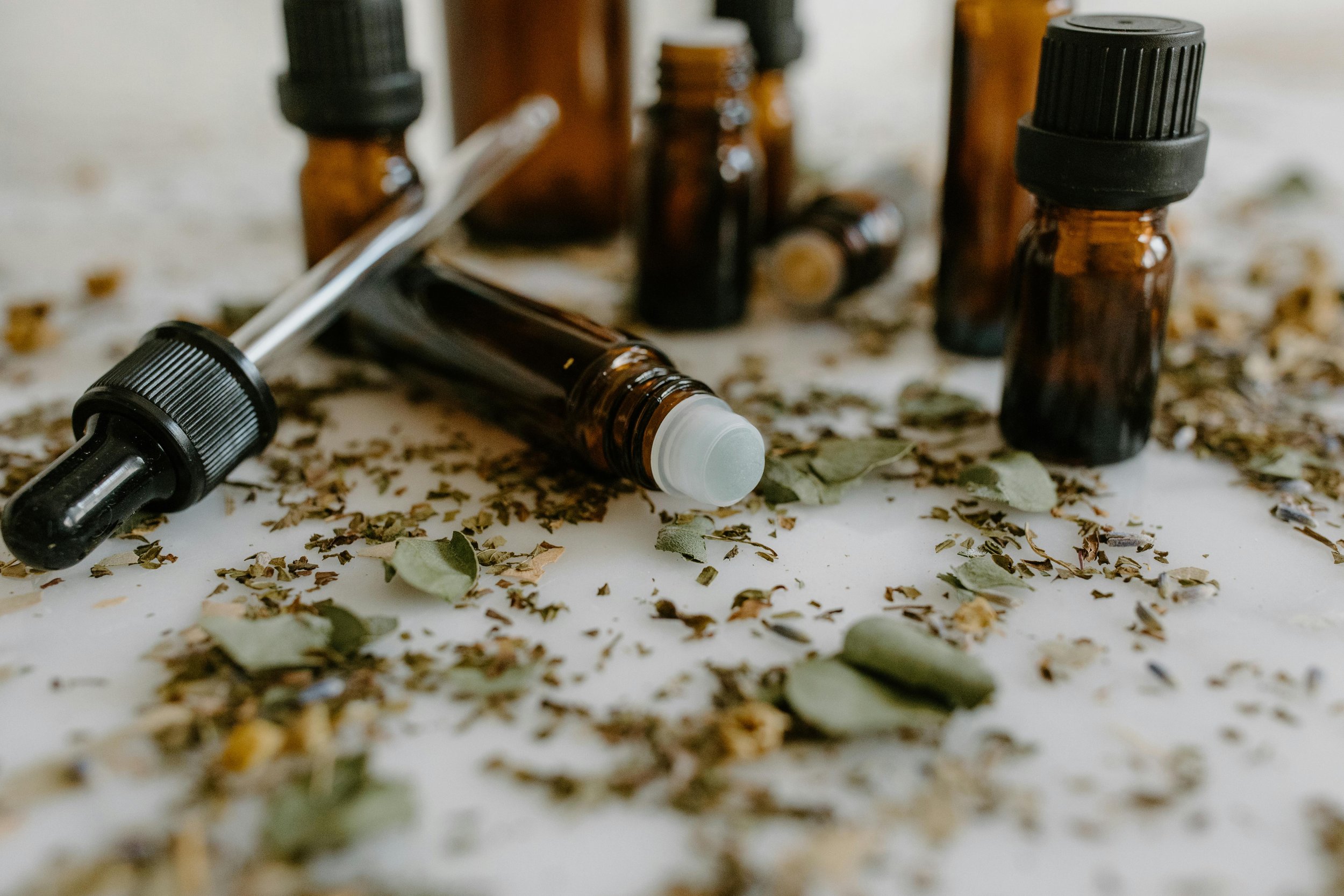Several small brown glass bottles and a dropper lying on a surface covered with dried herbs and leaves.