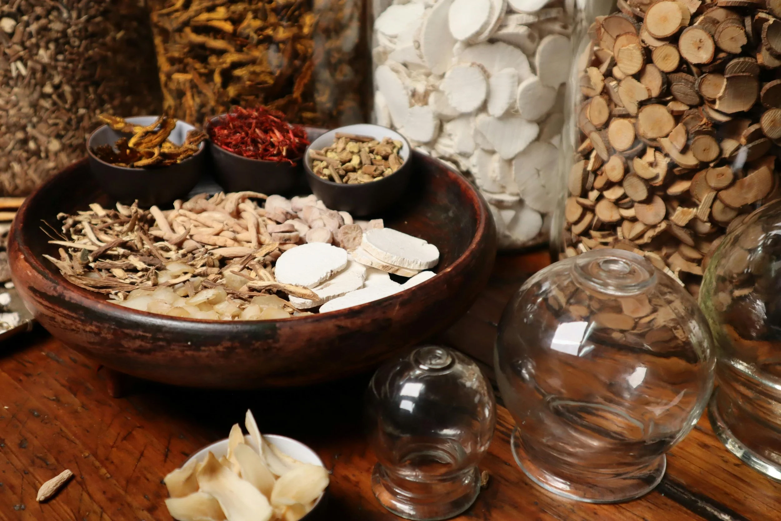 Various dried herbs, roots, and spices in small black bowls, along with white tablets and glass domes on a wooden surface.