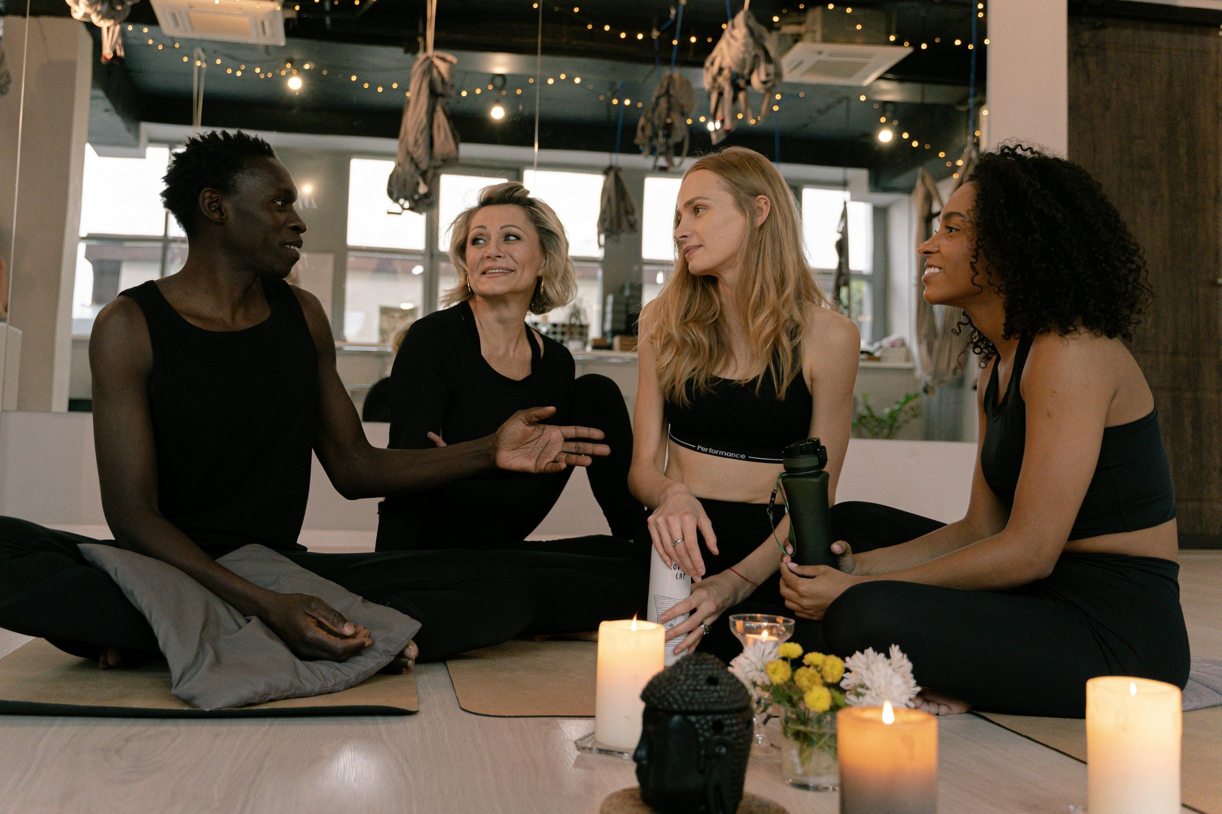 Four women sitting on mats in a peaceful room, engaged in a conversation, with candles and flowers on the floor.