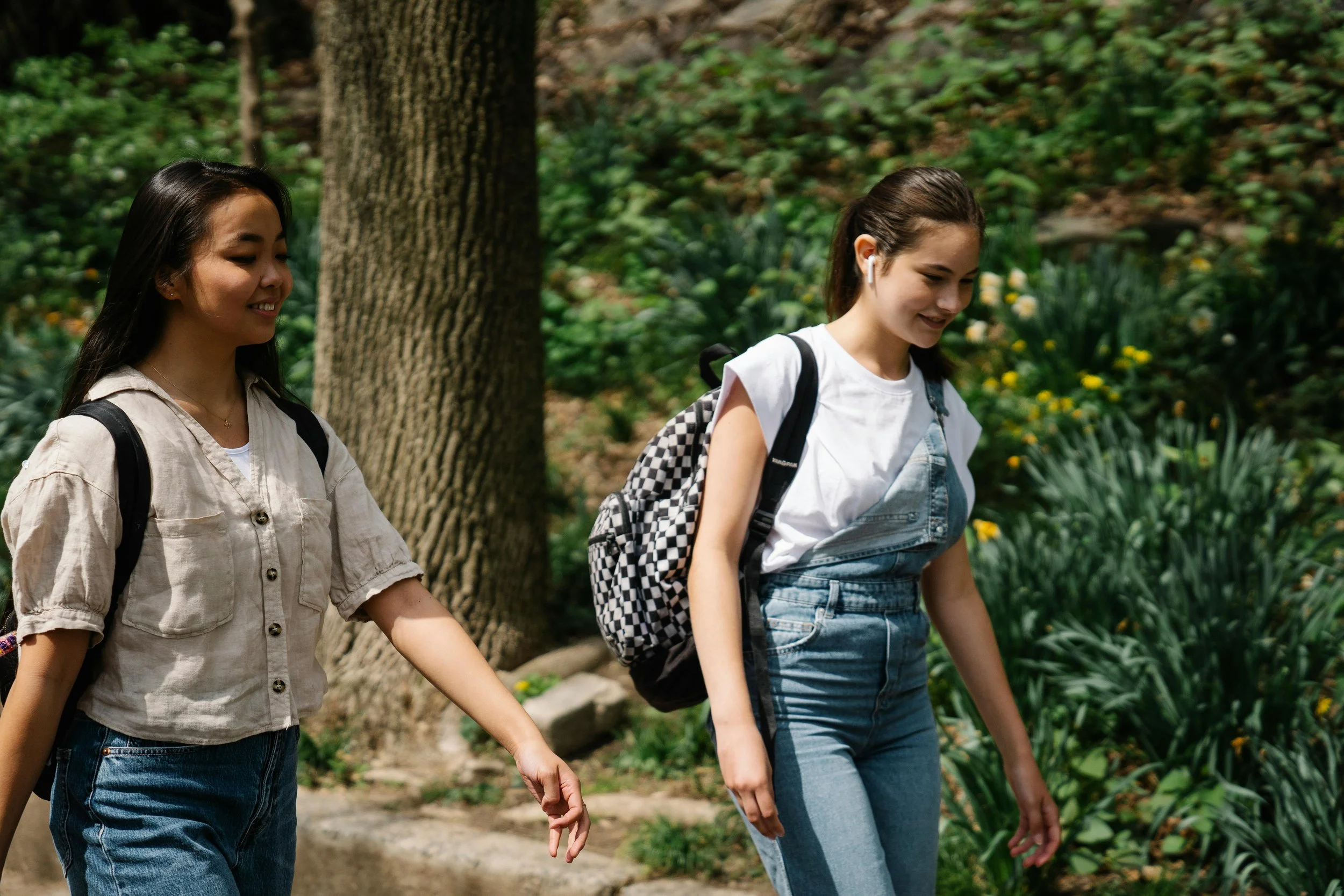 Two young women walking and smiling in a park, carrying backpacks, surrounded by trees and greenery.