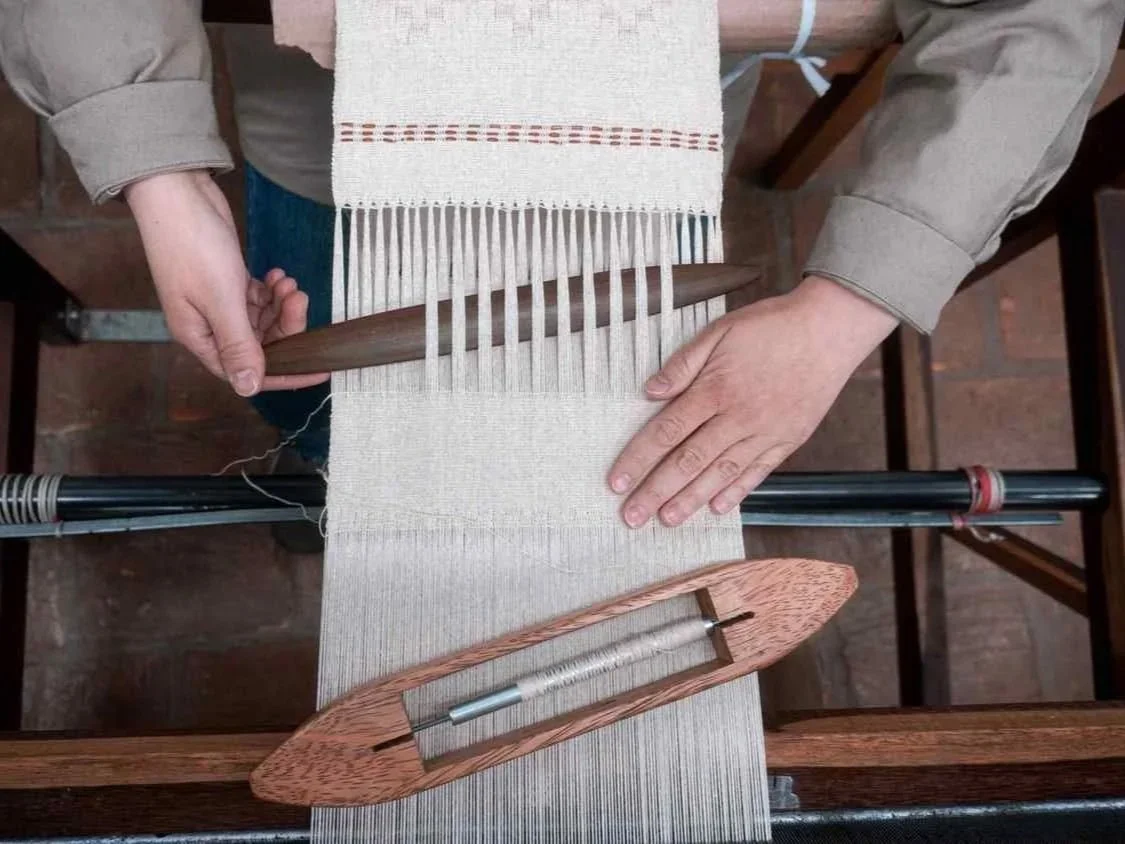 Person weaving on a traditional loom with a partially completed woven fabric.