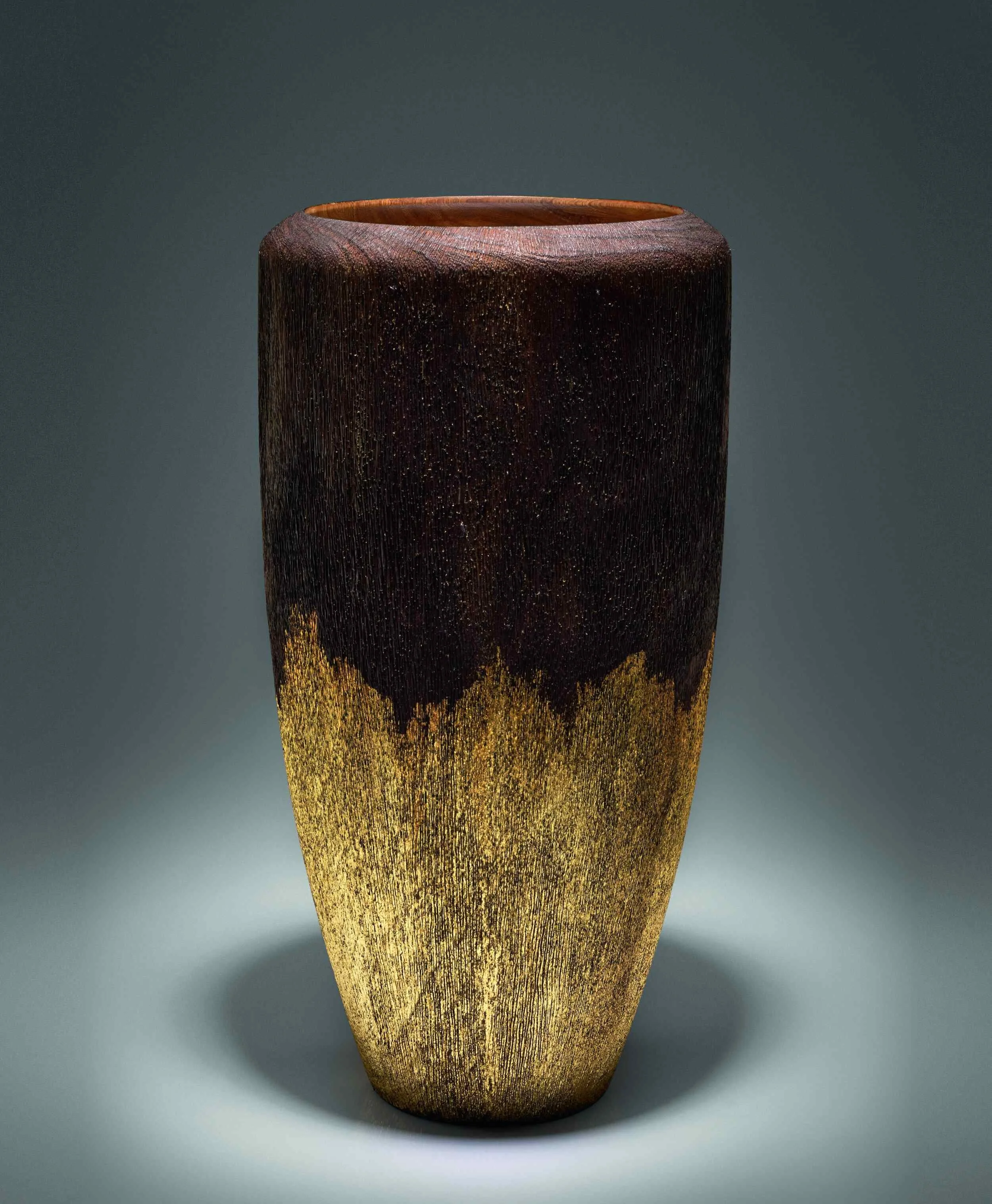A tall ceramic vase with a textured surface, transitioning from dark brown at the top to a goldish hue at the bottom, set against a plain gray background.