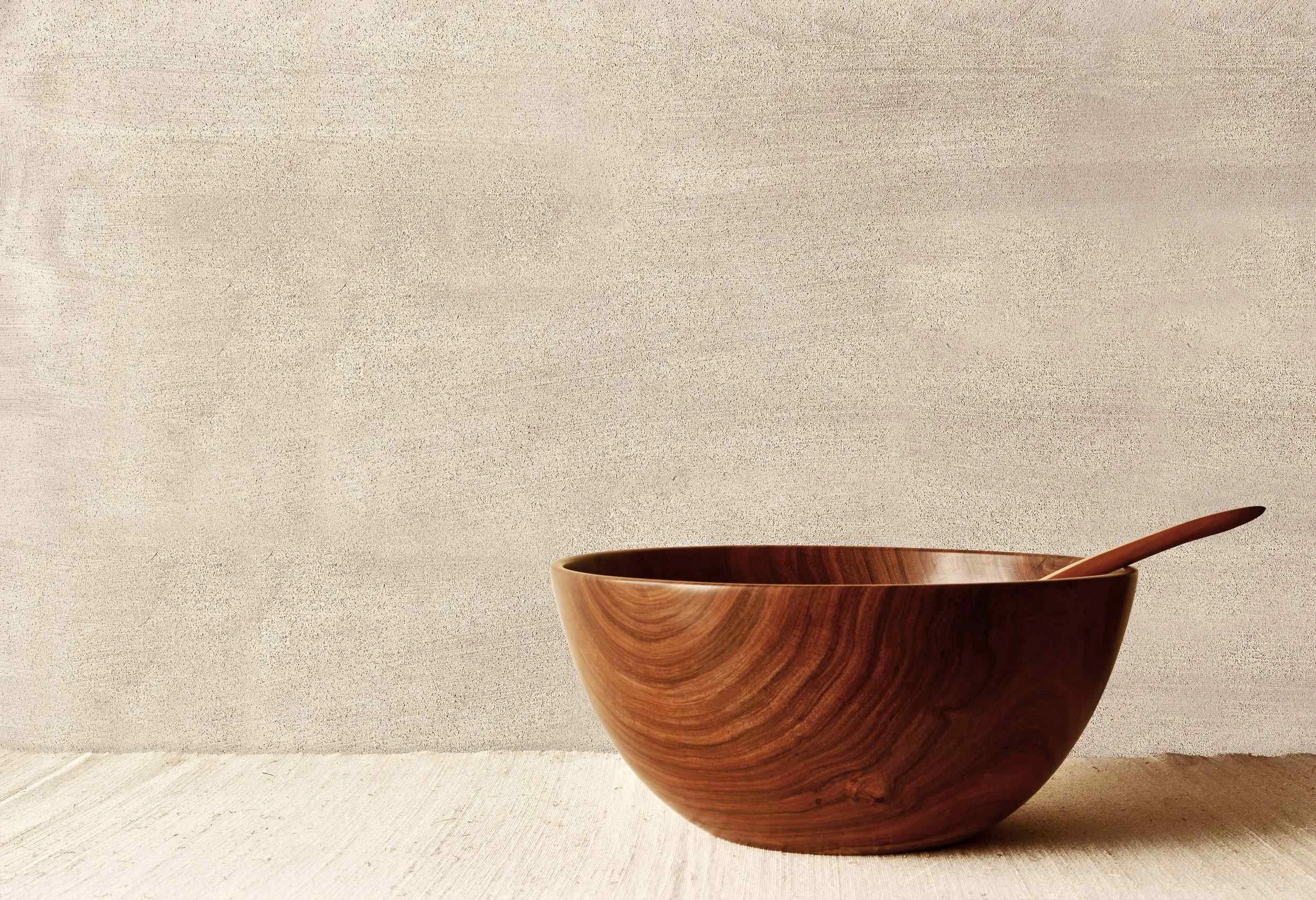 A wooden bowl with a matching wooden spoon on a light surface, against a plain, textured background.