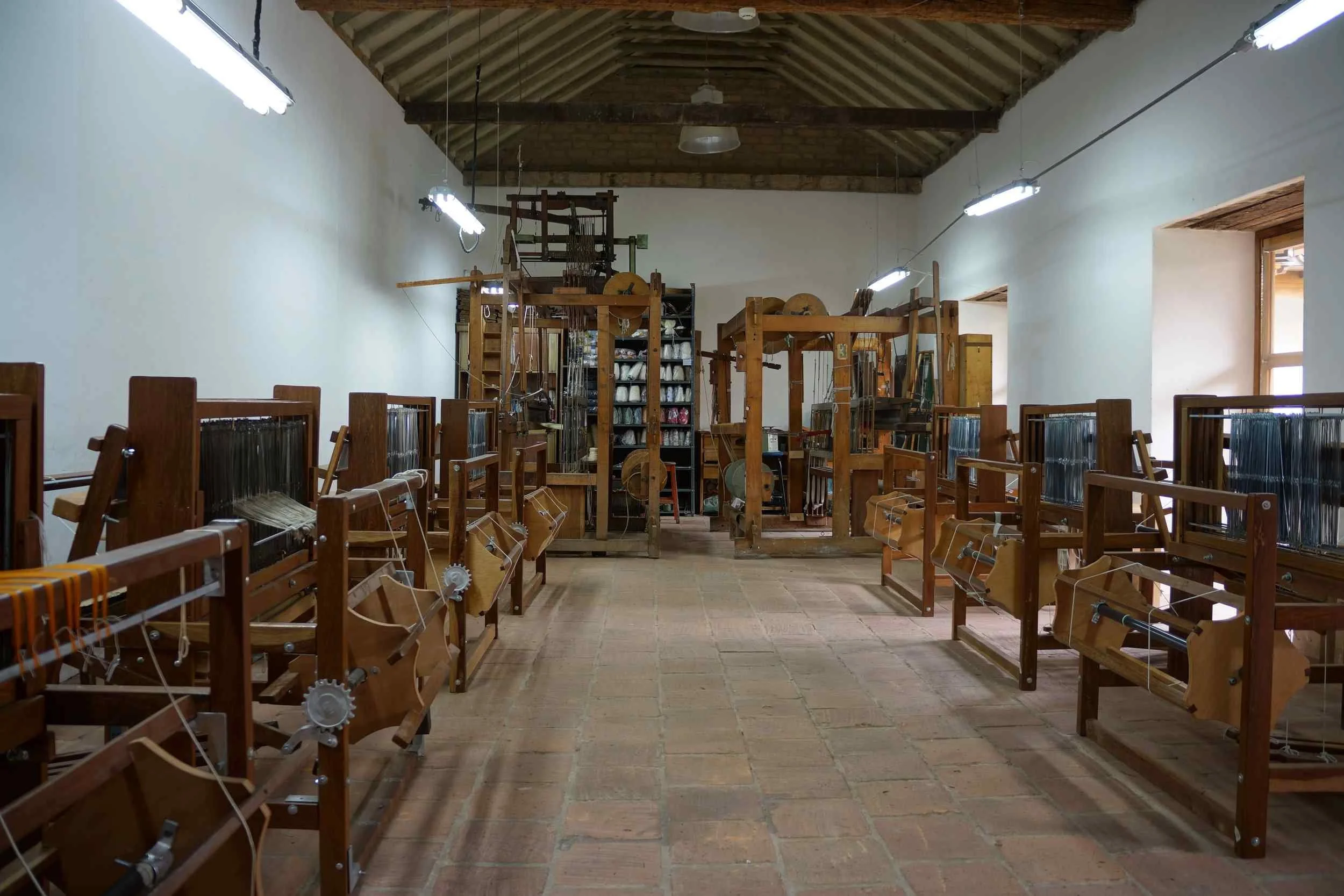 A weaving room filled with wooden weaving looms lined up on both sides of a brick floor and white walls, with large windows on the right side and ceiling lights above.